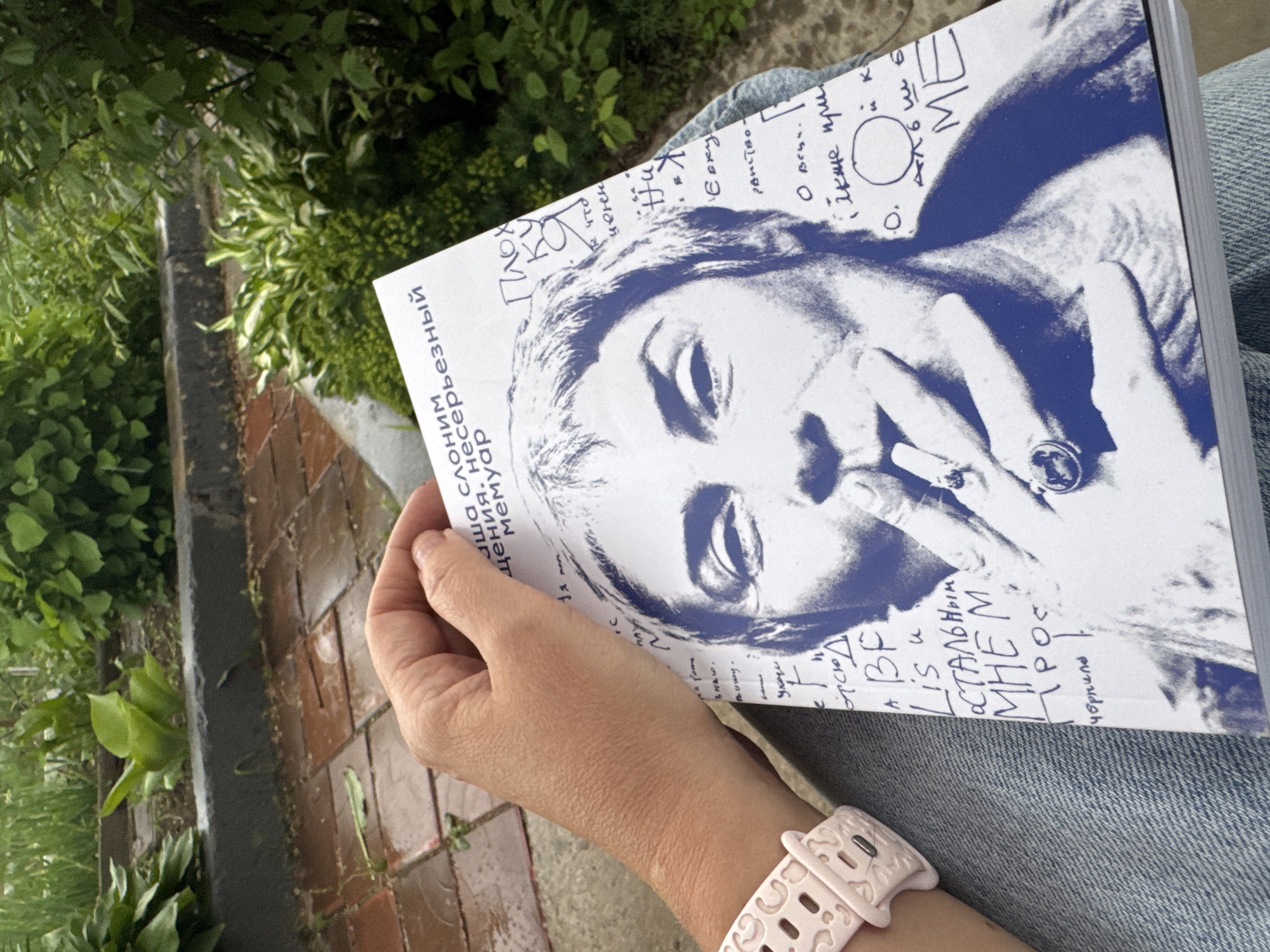 A person's arm and hand, adorned with a pink smart watch, hold a book with a striking blue-toned portrait of a woman smoking a cigarette. The book's cover features Russian text, some of which is obscured by the woman's face. The background consists of lush green foliage and a brick pathway, suggesting an outdoor setting. The image appears to be taken in a city park or garden.