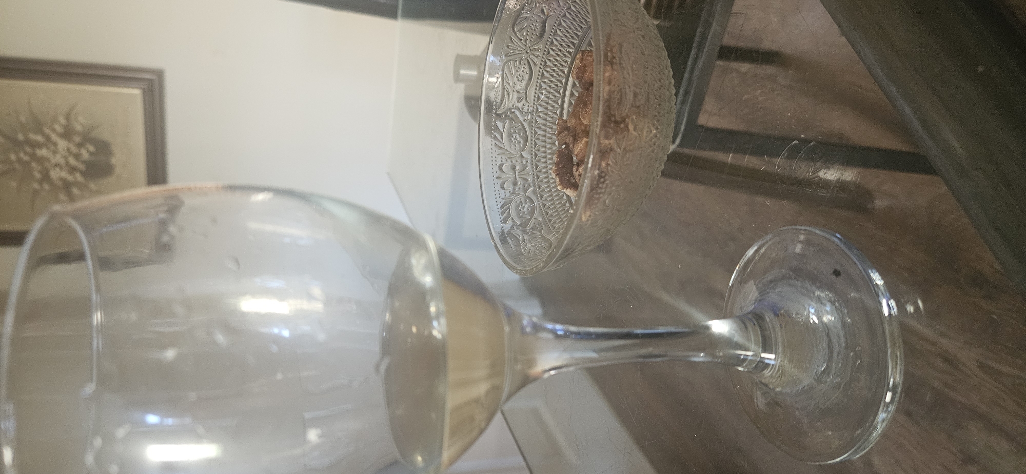 A wine glass lies horizontally on a reflective surface, likely a glass table, near a small, patterned glass bowl containing brownish, textured food items. In the background, a framed picture hangs on a wall. The reflective surface reveals elements of the room's furniture and decor. The location is unidentifiable as Norwich, United Kingdom based on the visible details in the image.