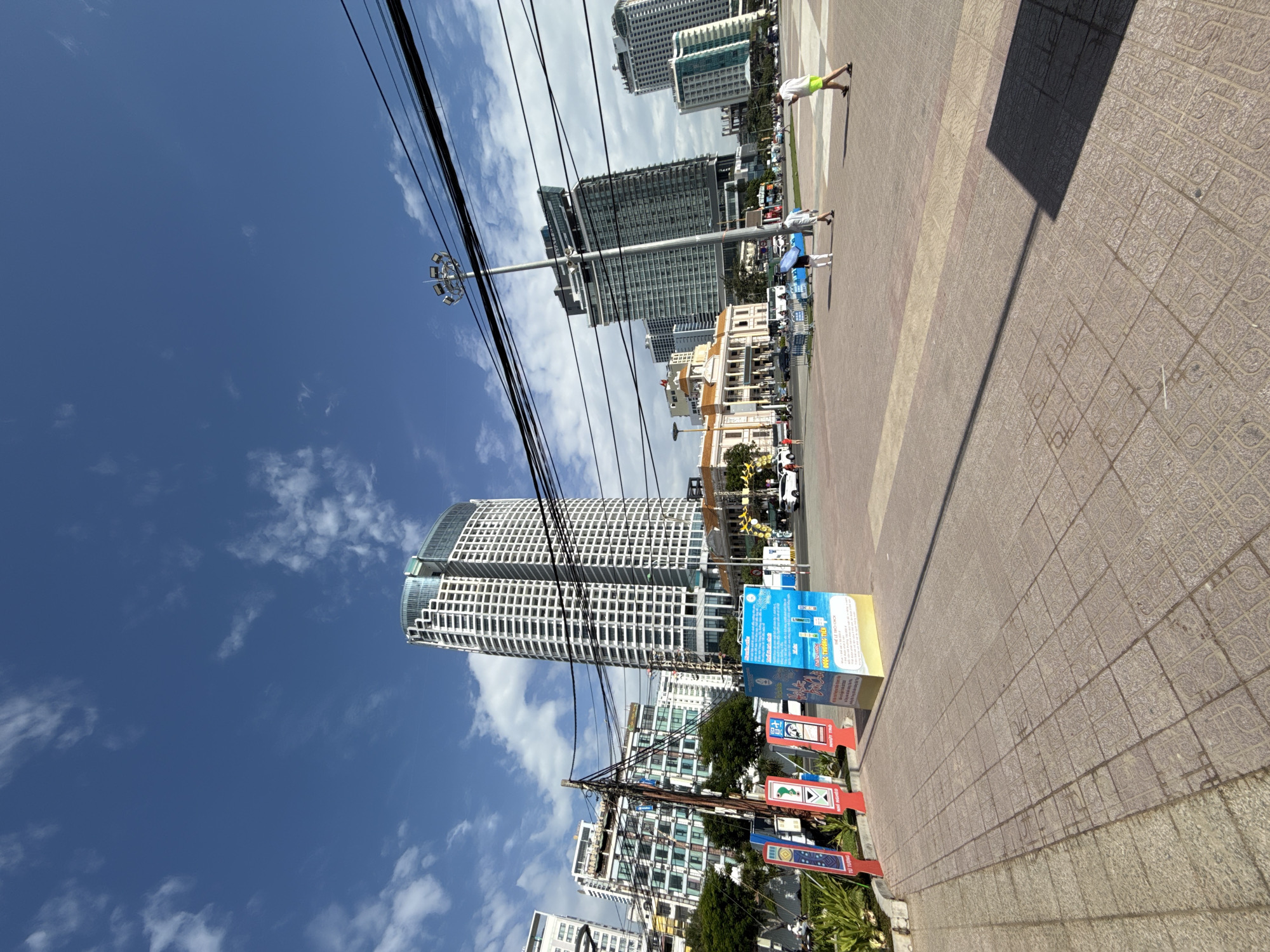 The image captures a sunlit urban scene in Nha Trang, Vietnam, under a bright blue sky with scattered white clouds. In the foreground, a tiled pedestrian walkway stretches towards the viewer, casting long shadows from unseen structures. A series of upright signs and a large informational billboard stand to the left of the walkway. Beyond this, a wide street is visible, populated by vehicles including cars and a bus.
Dominating the mid-ground and background are several tall, modern buildings, characteristic of a city skyline. The most prominent building is a slender, white skyscraper with a distinctive curved top and a grid-like facade. To its left and right, other high-rise structures of varying architectural styles are present, some featuring glass facades and balconies. A large, light-colored building with a colonial architectural style, featuring arched windows and columns, is situated near the center of the mid-ground.
Several people are visible in the scene. To the right of the walkway, a man in a yellow shirt and green shorts walks away from the camera, holding a phone. In the middle of the street, two individuals are walking, one holding a blue umbrella, and another person is seen in the distance to the far left. Overhead, a dense network of power lines crisscrosses the sky, supported by utility poles. The overall impression is of a vibrant, bustling city environment on a clear day.