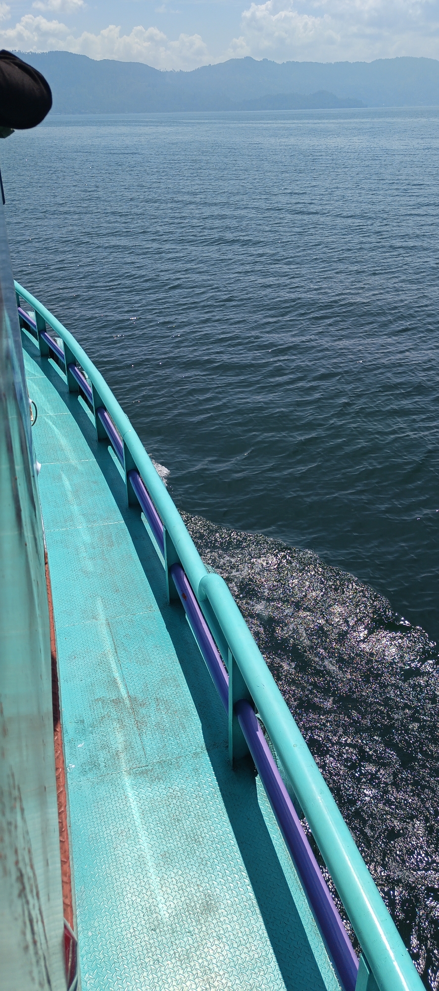 The image captures a perspective from the deck of a boat traveling on a body of water. In the foreground, a turquoise deck with a diamond plate pattern is visible, leading towards the right edge of the frame. A railing system, painted turquoise with purple bars, runs along the edge of the deck. Beyond the railing, dark blue water with ripples and white foam from the boat's wake is evident. In the distance, a hazy blue mountain range borders the horizon under a partly cloudy sky. The specific location in Cinta Dame, Indonesia is not visually discernable from the image itself. There are no people visible in the frame.