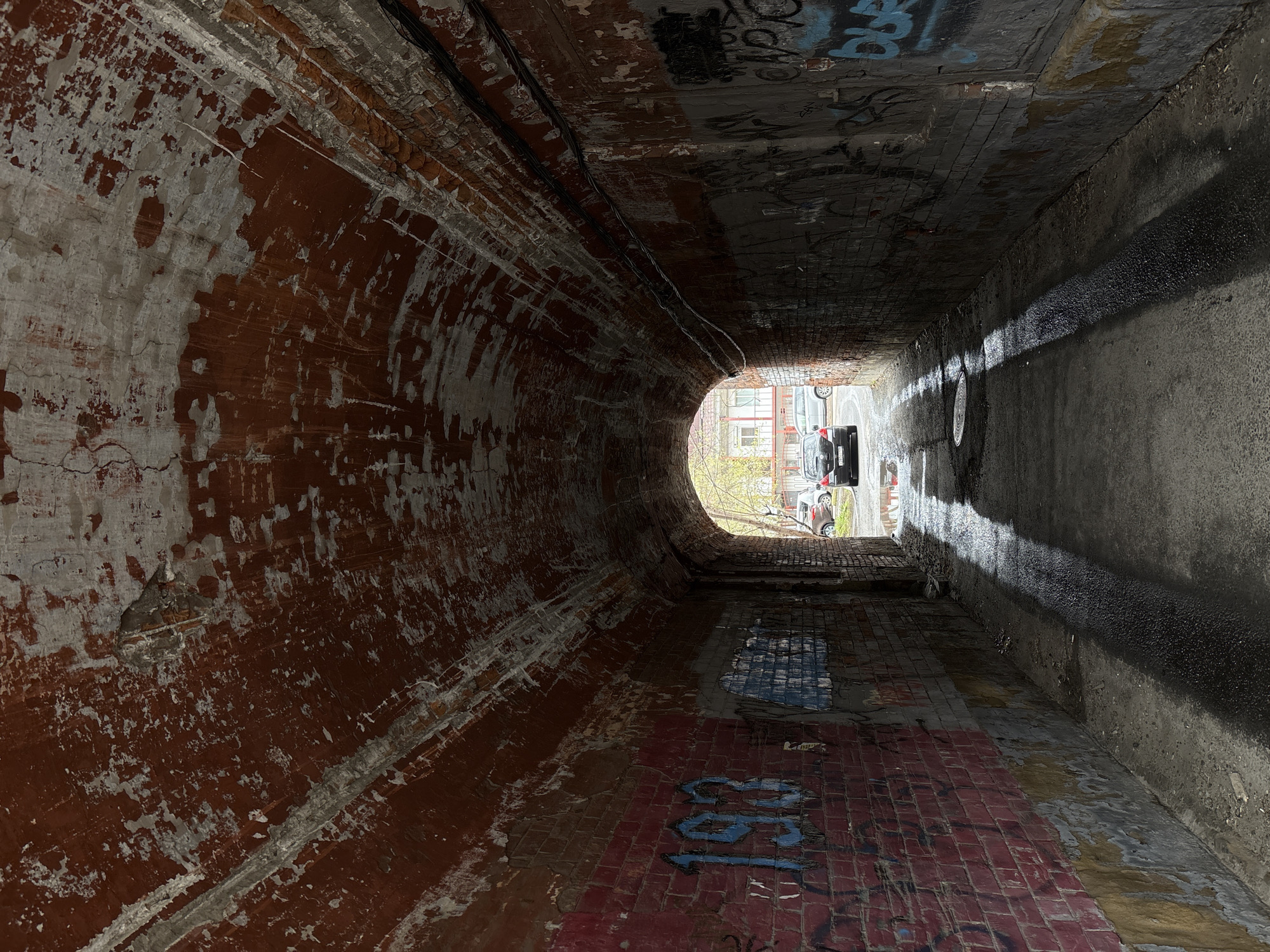 The image depicts the interior of a pedestrian tunnel in Irkutsk, Russia. The tunnel's walls are curved and constructed of brick covered with graffiti and decaying paint. The left wall displays a faded red undercoat beneath peeling white paint. The right wall features more modern graffiti and appears wet with pooled water. The tunnel floor is partially tiled and similarly marked with graffiti. At the tunnel's exit, sunlight illuminates the street beyond, revealing a parked car. The tunnel environment is dark and appears poorly maintained.