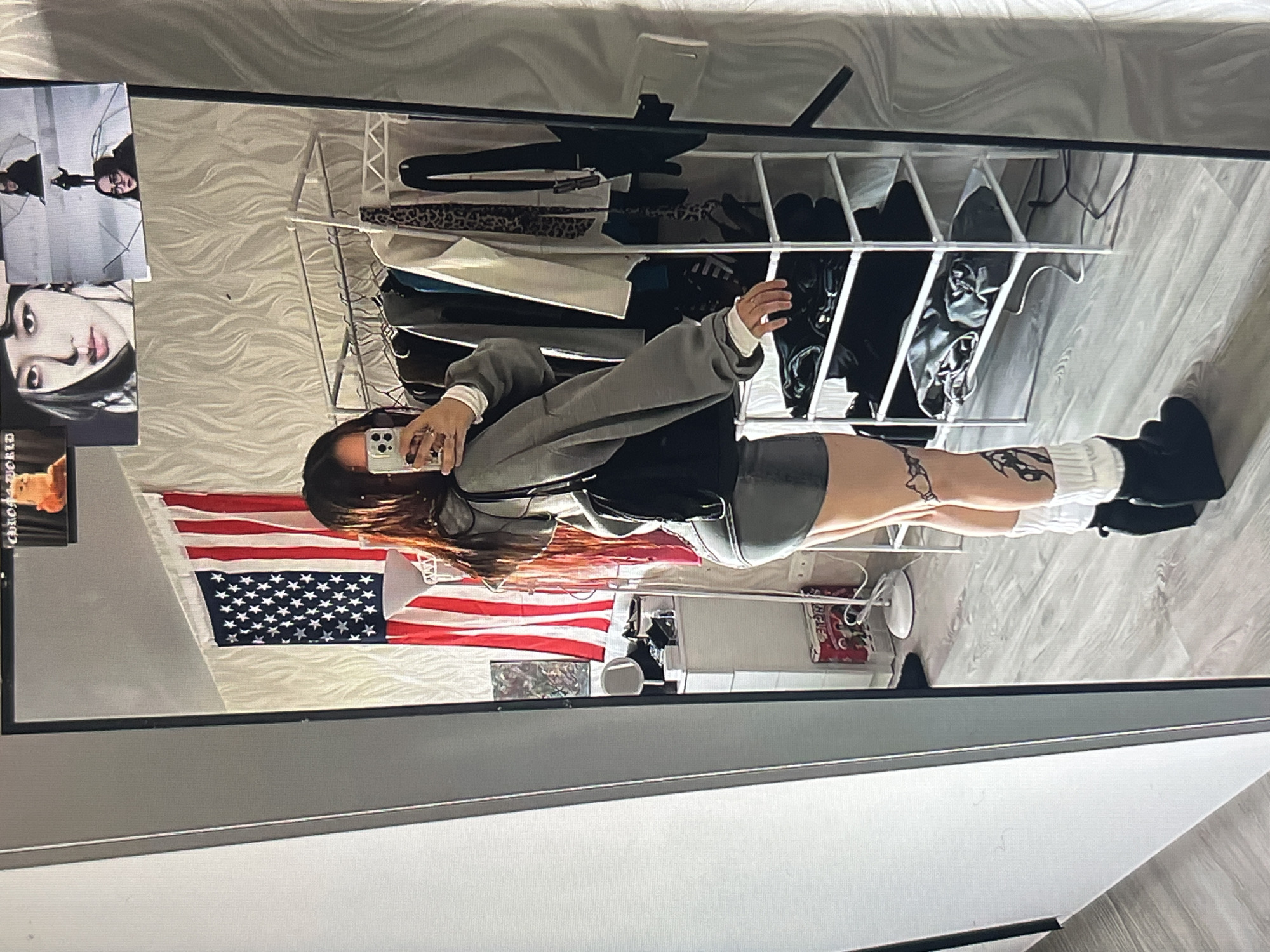 A young woman is taking a mirror selfie in what appears to be a bedroom. She is wearing a grey oversized sweatshirt, a grey mini skirt, white leg warmers, and black boots. She has long, dark hair and visible tattoos on her legs. Behind her, an American flag is hanging on the wall. There is a clothing rack with various items of clothing and a white shelving unit. A small table with a lamp and some drawers is also visible. In the mirror, there are a few framed pictures on the wall above the flag, including one of a woman's face and another with a landscape. The floor appears to be laminate flooring. There is no indication that this image is located in Krasnoyarsk, Russia.