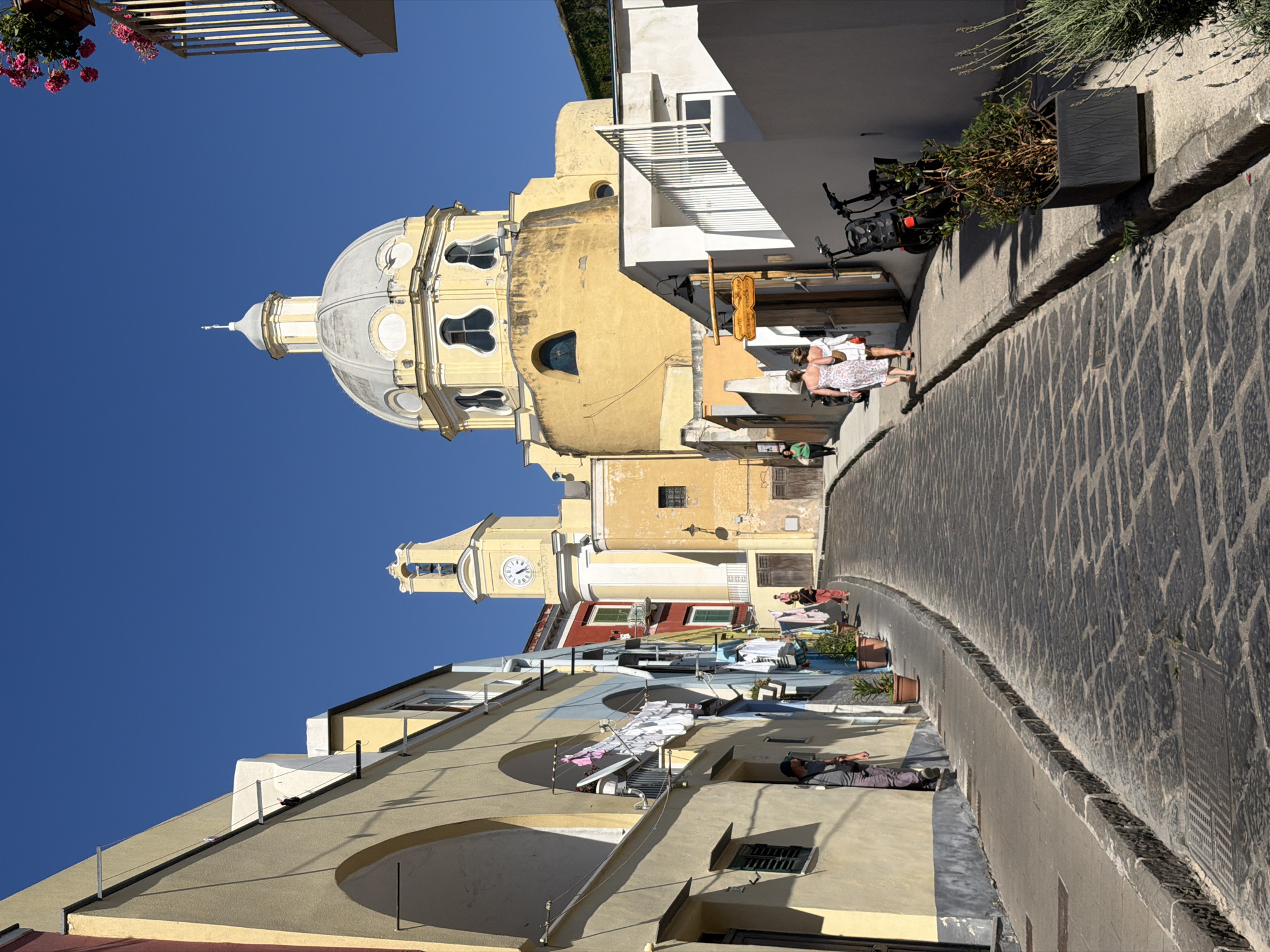 A vertical view of a street in Procida, Italy, is shown with buildings and the dome of the Church of Santa Maria delle Grazie Incoronata. The street is paved with dark gray cobblestones, and runs from lower right to upper left. A retaining wall is visible along the right side of the street, covered with a plant.
Along the left side, buildings are stacked closely together, with white clothes hanging across some of the windows and balconies. A man is laying on a rooftop on the left side. Two women are standing and talking along the road. A yellow church with a gray dome is on the left side of the street. Above the church is a clear blue sky. A basket of pink flowers is hanging above.