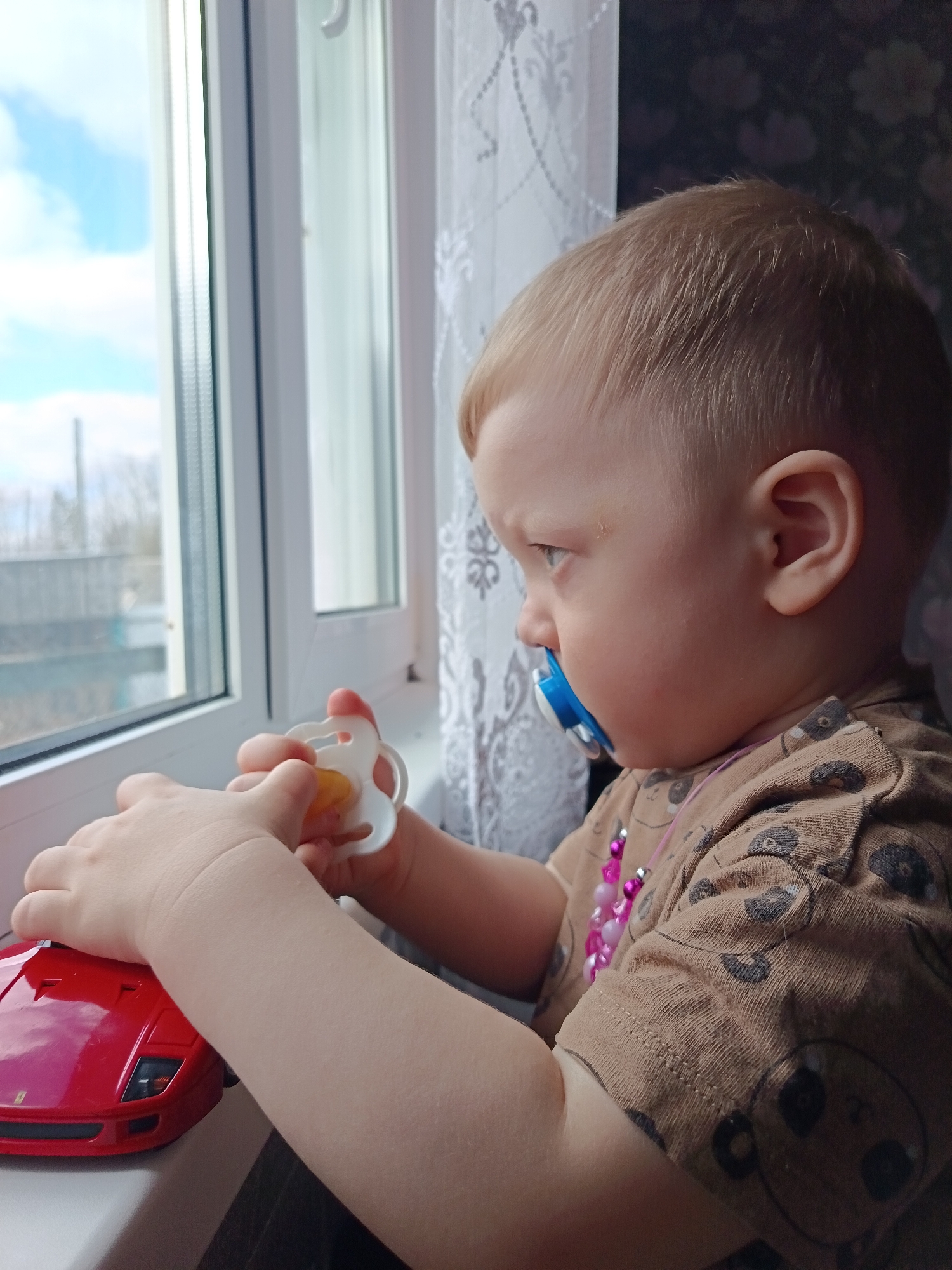 A young child with light blonde hair, wearing a brown shirt with a monkey pattern and a pink beaded necklace, stands on a windowsill, gazing out the window. The child has a blue pacifier in their mouth and is holding another pacifier in their left hand. A red toy car sits on the windowsill near the child's hands. Outside the window, a cloudy sky is visible above a landscape with trees and buildings. The backdrop includes patterned wallpaper. The location is Nizhnyaya Chermoda, Russia.