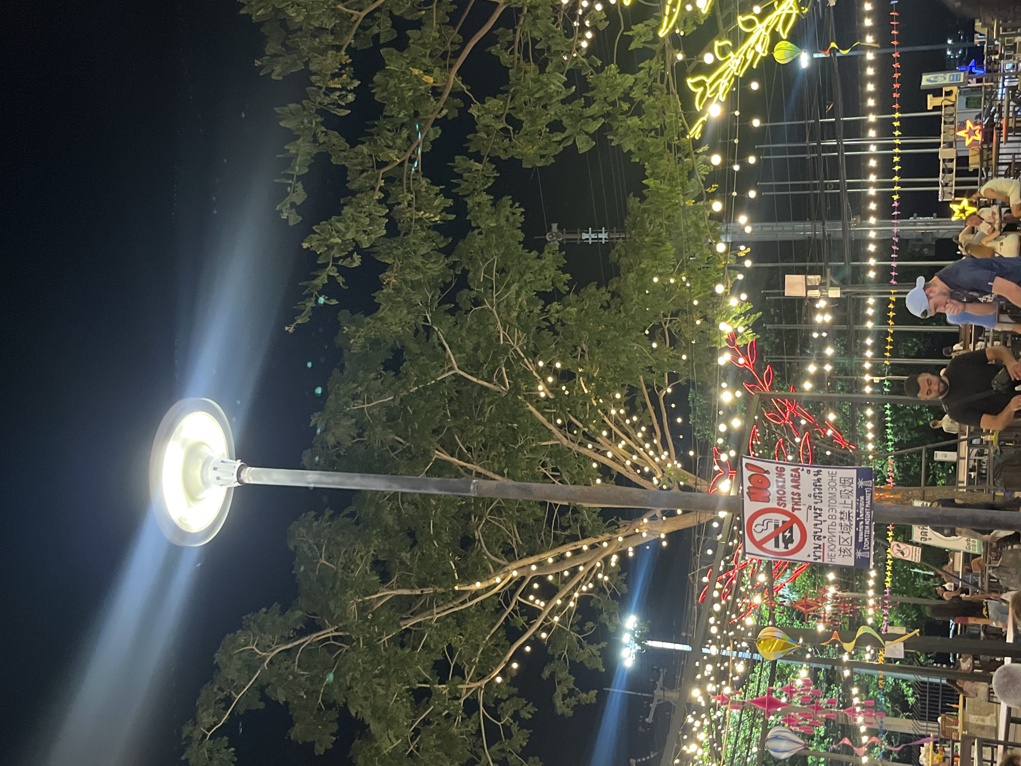 The image depicts a night scene in Pattaya City, Thailand, likely at the Jomtien Night Market, as indicated by a sign. A prominent feature is a street lamp shining brightly on the left. A tree with small decorative lights interspersed among its branches occupies the center of the frame, with some of the branches going over the street lamp. Other decorative lights stretch across the scene, including neon lights in leaf designs. A "No Smoking" sign in multiple languages is affixed to a pole. In the background, one sees people dining in the market. There is also the presence of decorations, such as colorful kites.