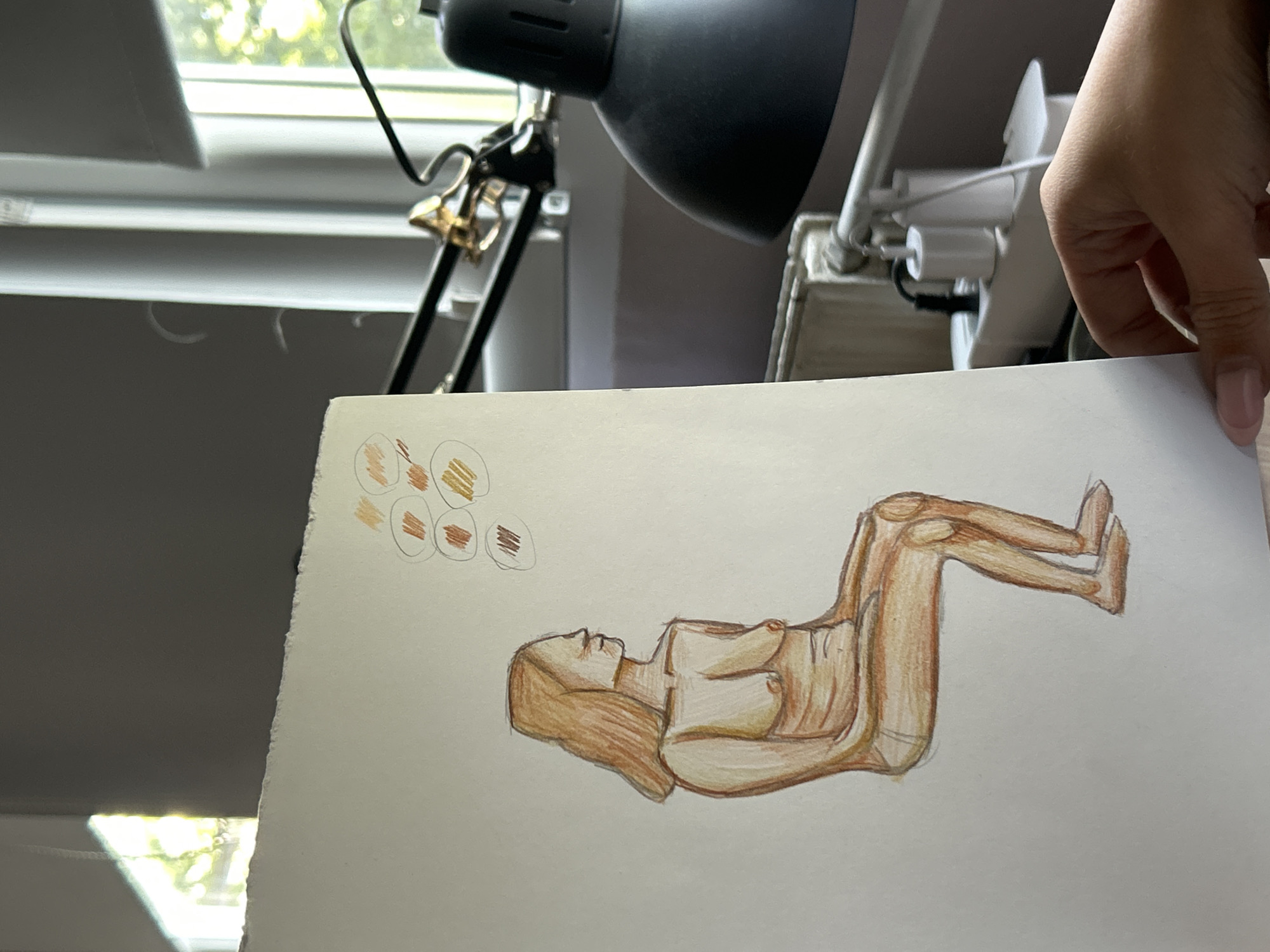 A hand holds a sheet of paper with a drawing of a reclining nude figure rendered in shades of brown and beige. The drawing also features color swatches near the top left corner. The background includes a black adjustable desk lamp, a white window blind, and a glimpse of greenery outside the window. The location is not discernible from the image and cannot be pinpointed to Sofia, Bulgaria.