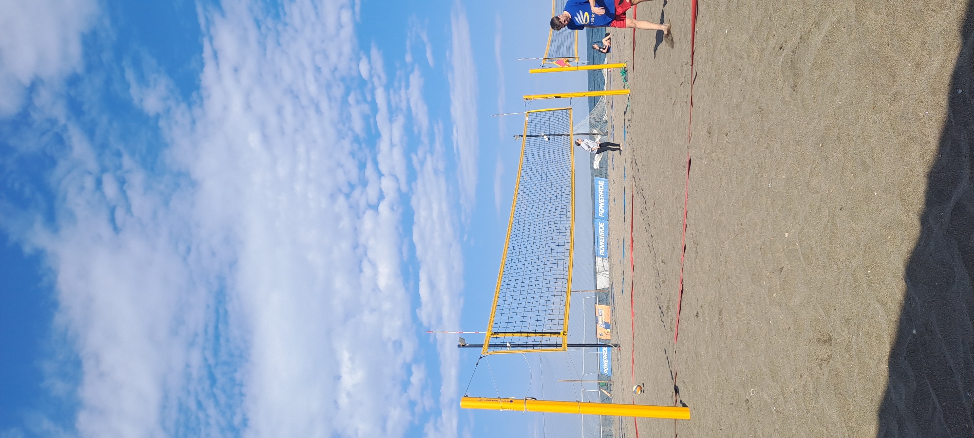 This image captures a vibrant beach volleyball scene under a bright, partly cloudy sky in Burgas, Bulgaria. The sky is a clear blue with scattered white, wispy clouds, suggesting fair weather.
The primary subject is a beach volleyball court made of light-colored sand, marked with red boundary lines. Two yellow-framed volleyball nets stretch across the sand, with the closest one prominently featured in the foreground. One orange and white volleyball rests on the sand near the base of the closest net's left post. Yellow net posts support the nets, with a clear blue ocean visible in the background.
Several people are present on the courts. In the foreground, a person wearing a blue shirt with "CURRY" printed on it and red shorts stands barefoot on the right side of the closest court, seemingly observing or preparing to play. Further back, beyond the closest net, another person is seen sitting or lying on the sand, while a third person in dark pants and a light top stands near a more distant net.
The time of day appears to be either mid-morning or late afternoon, indicated by the strong sunlight and visible shadows cast on the sand, including a long shadow on the far right. Blue banners displaying "POWERADE" vertically are visible along the side of the court, acting as sponsor signage or court dividers. A faint website address, "www.volleyplay.com," can be seen on the yellow base of one net post. The overall atmosphere suggests a casual or recreational volleyball event.