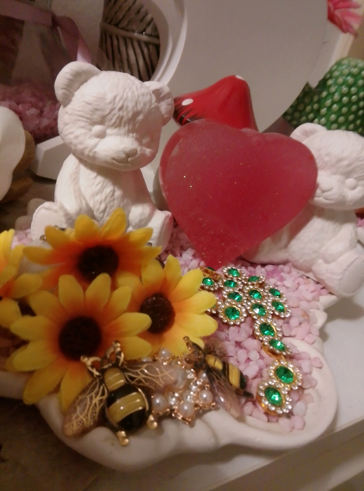 The image shows a close-up view of a decorative arrangement, likely a craft project or a display. In the foreground, there are several artificial yellow sunflowers with dark centers. Attached to the sunflowers and interspersed with small pink pebbles are various decorative charms. These include two bee charms with black and yellow stripes and gold accents, a gold cross encrusted with green gemstones, and a cluster of small white pearls. Behind the flowers and charms, a large, translucent red heart-shaped object with glitter is prominently displayed. To the left of the heart is a white ceramic bear figurine, facing slightly forward. To the right of the heart is another white ceramic bear figurine, partially visible and facing to the right. The background is blurred but suggests other decorative items, including a hint of a red mushroom shape. The overall impression is a whimsical and colorful collection of decorative objects. The image was taken in Wedel, Germany.