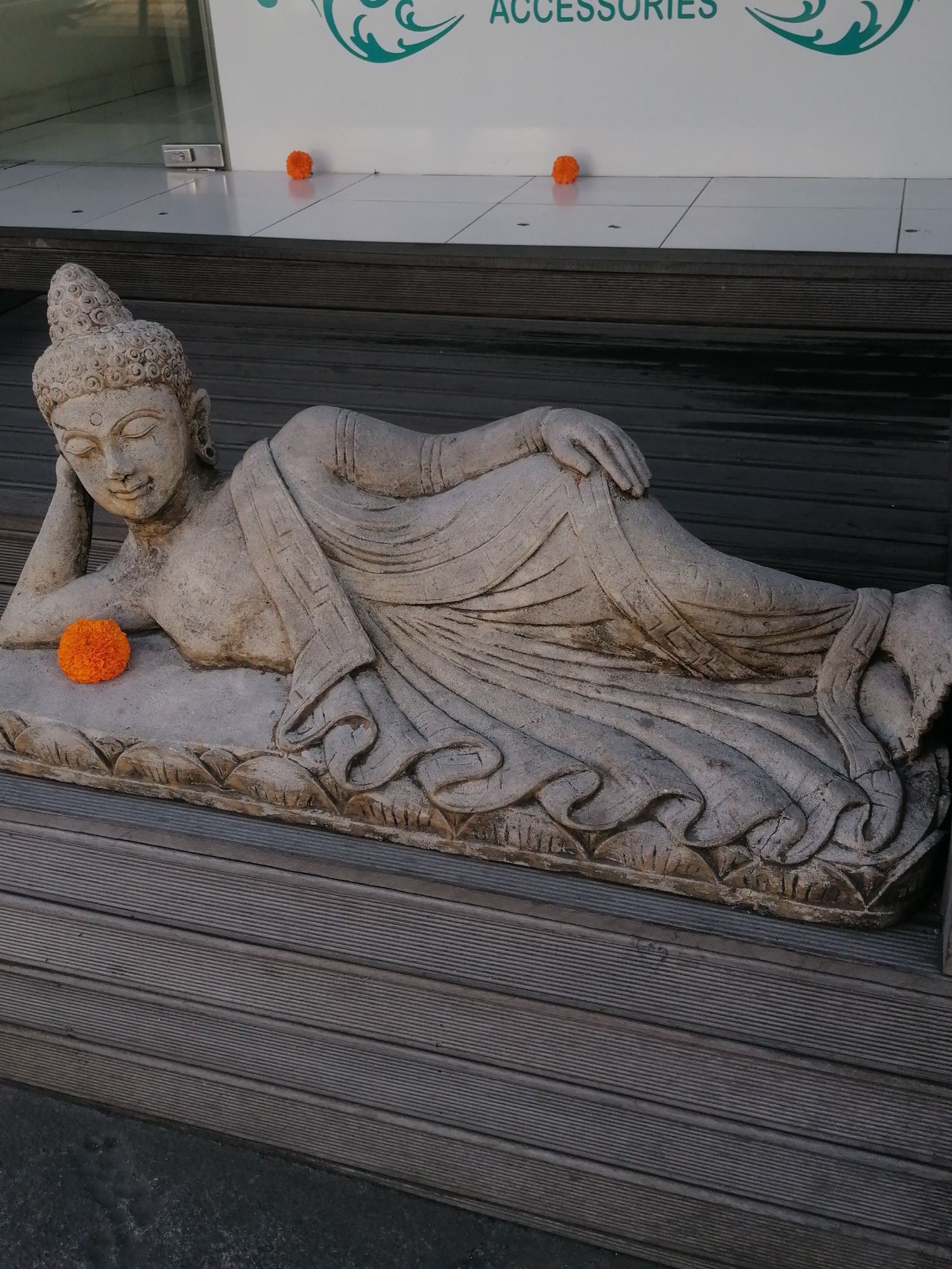 A stone statue of a reclining Buddha rests on a wooden deck. The Buddha is depicted with its head resting on its arm, eyes closed, and draped in robes. An orange marigold flower is placed on the statue's chest. The statue lies in front of a building with a white facade featuring a green decorative motif and text that appears to read "ACCESSORIES". Two similar orange flowers are visible on the tiled floor in front of the building. The scene is located in the city of Ubud, Indonesia.