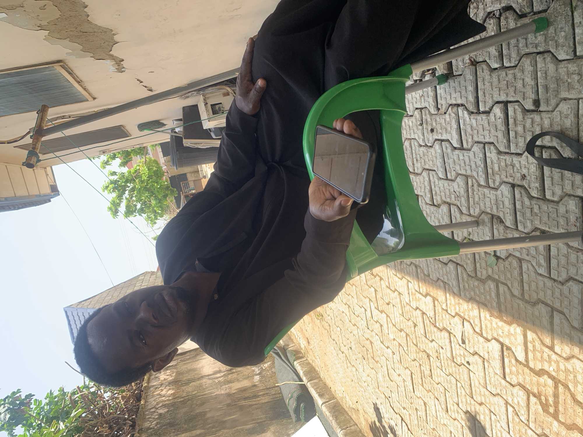 A Black man, wearing a black traditional outfit and sitting on a green plastic chair, is holding a smartphone in his right hand. The man is positioned outdoors, with a paved area of interlocking concrete bricks in the foreground. Sunlight casts shadows on the paving. In the background, there is a building with a light-colored, partially damaged facade, and some greenery. The scene appears to be in a residential or commercial area, potentially in Abuja, Nigeria.