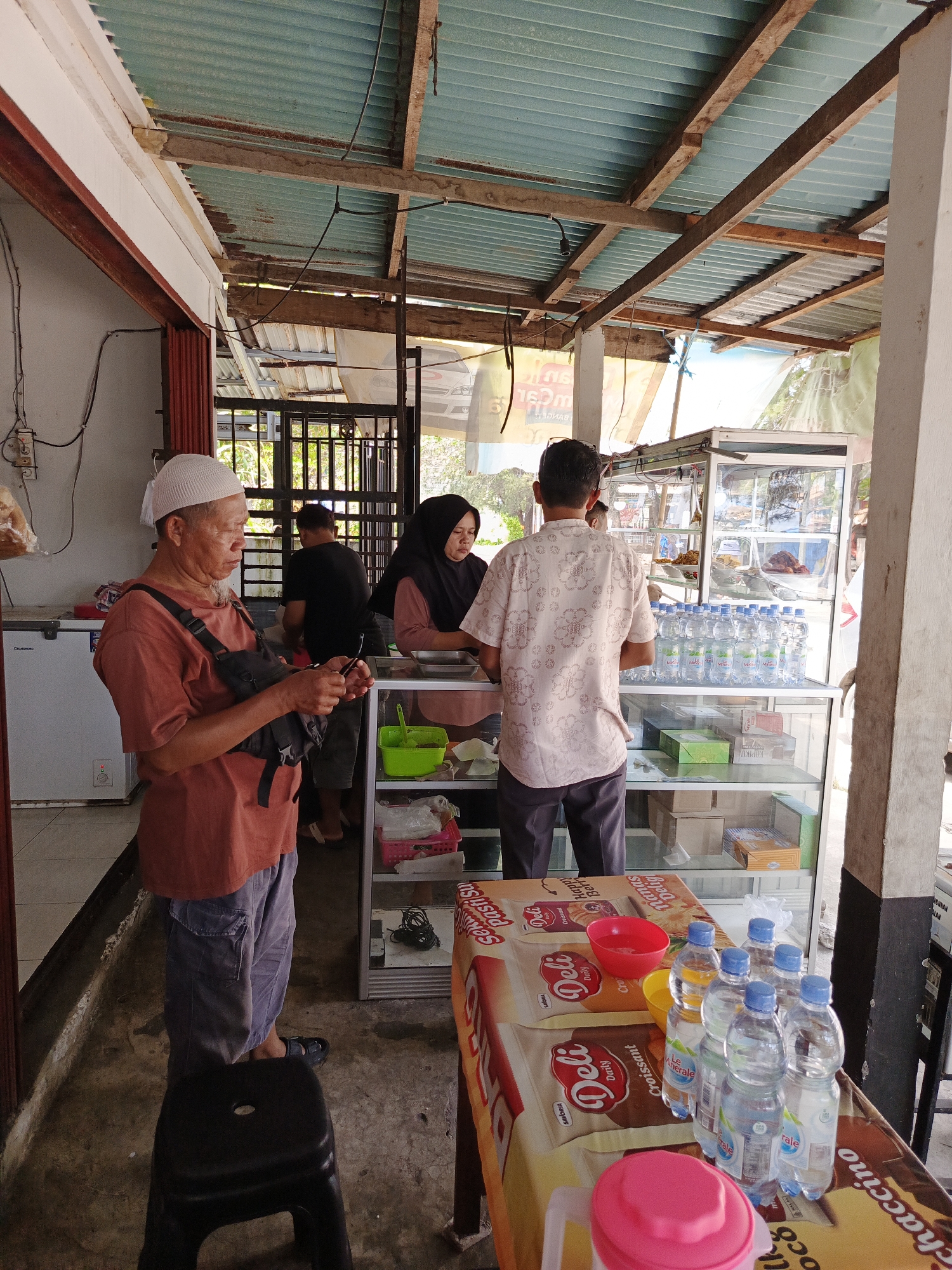 In a roadside food stall in Bengkulu, Indonesia, an elderly man wearing a white cap and a terracotta-colored shirt, with a black bag slung across his chest, stands holding a smartphone. To his right, a woman in a black hijab and pink shirt attends to customers. Behind the counter, a man in a patterned light-colored shirt faces away from the camera. The stall features a glass display cabinet filled with bottled water, boxed snacks, and some food items. A red bowl and a yellow bowl are visible on a table covered with a colorful cloth featuring food advertisements. The roof of the stall is constructed from corrugated metal supported by wooden beams, and the overall environment appears open-air.
