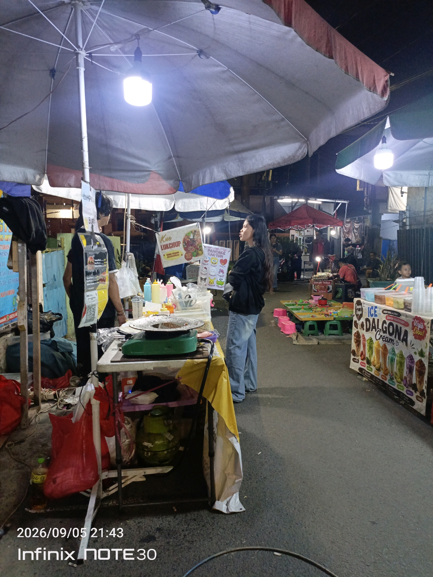 The image shows a street food stall at night in an outdoor market. A young woman in a black hoodie and blue jeans stands near a food preparation area. The stall has a large umbrella providing shade, with a bright light hanging from it. There are various food items and ingredients on display, including colorful drinks and a grill. Several other people are visible in the background, some walking and others at different stalls. The ground is paved, and there are other market stalls and tents in the distance, indicating a busy commercial area. The overall scene suggests a lively evening market atmosphere.