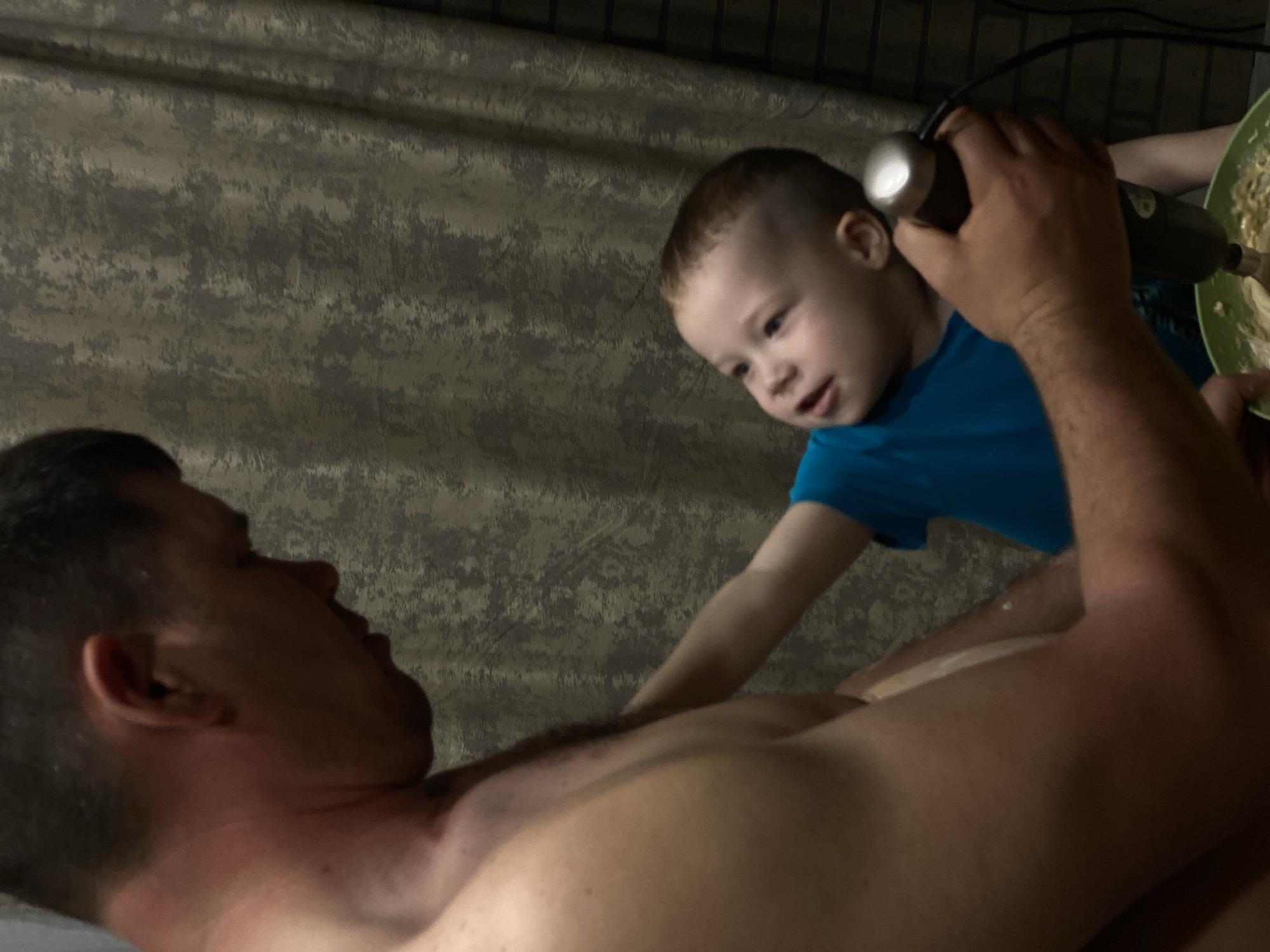A partially nude adult male lies horizontally facing left, while a young child in a blue shirt stands beside him, holding an immersion blender over a bowl. The man's upper torso and the child's arm are visible. The background appears to be a textured wall. There's no evidence indicating the location is Sochi, Russia.
