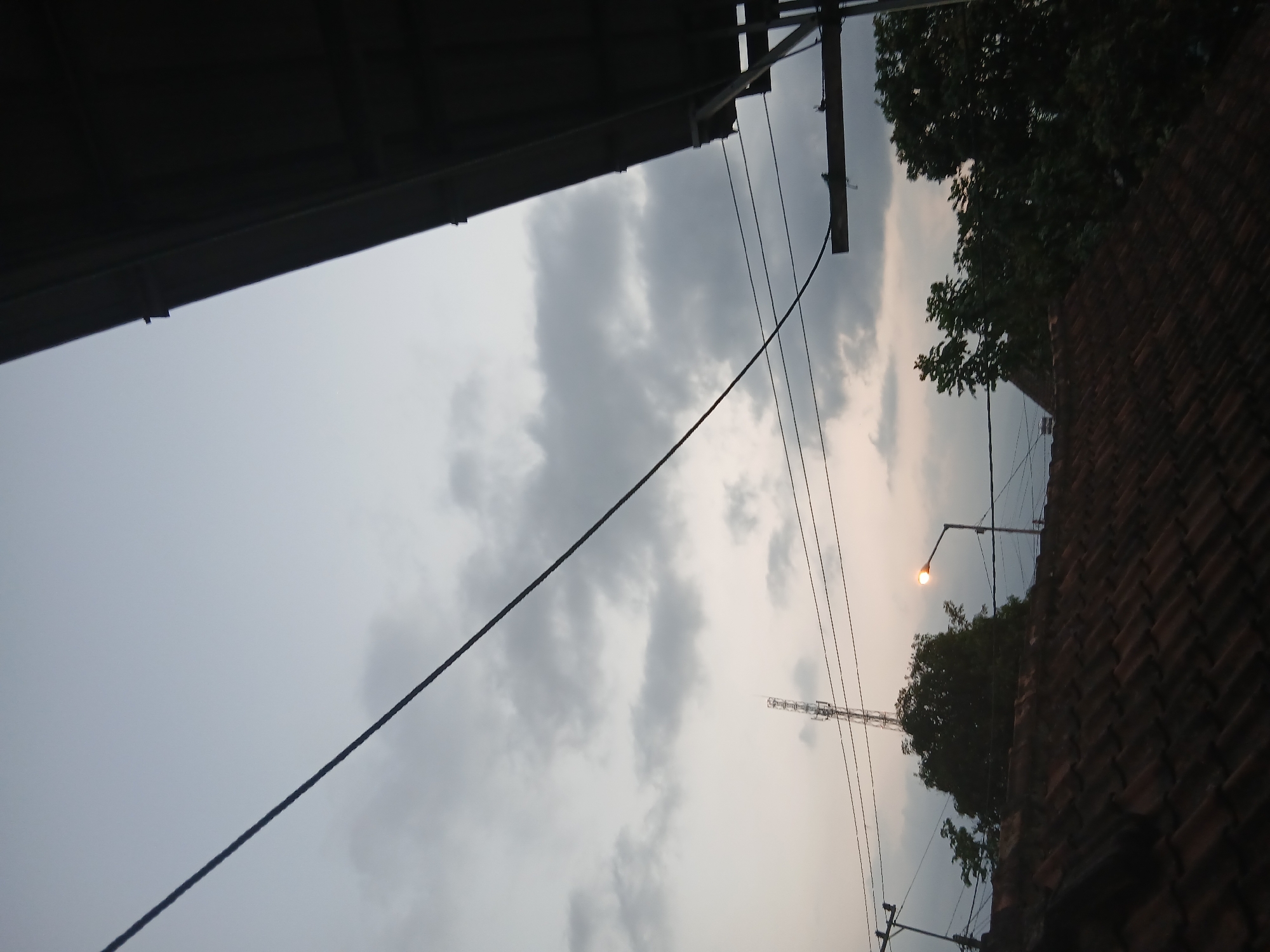 The image depicts a sky view in Kademangan Kulon, Indonesia, likely during dusk or dawn due to the diffused light and overcast conditions. The foreground is dominated by a tiled roof of a building, suggesting a residential or urban setting. Above the roofline, a transmission tower is visible in the distance, indicating the presence of communication infrastructure within the city. Several power lines stretch across the frame, crisscrossing the sky. A street lamp with its light on is also present, casting a warm glow. Lush green trees are scattered across the landscape, interspersed with structures, suggesting a blend of natural and built environments. The overall atmosphere is subdued, with the cloudy sky creating a soft, diffused light.