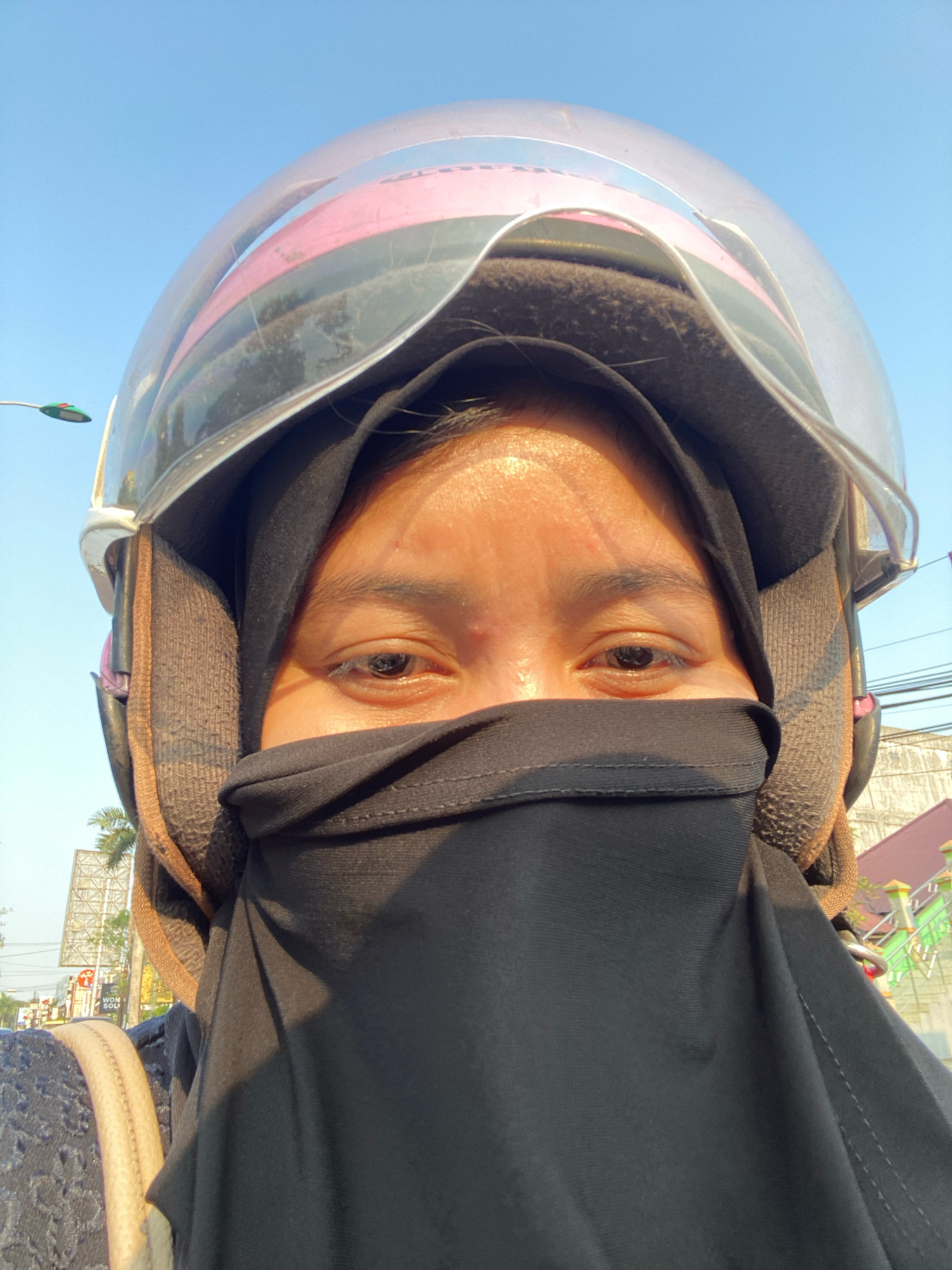 A close-up, front-facing selfie shows a person wearing a black hijab and a black face covering, obscuring most of their face. The visible part of their face reveals one eye, with an eyebrow and forehead visible above. They are wearing a helmet, with the clear visor partially raised, revealing the top portion of the helmet's interior padding and a pink accent. The background suggests an outdoor urban environment with a clear blue sky. In the far background, there are hints of buildings, power lines, and greenery, possibly palm trees, indicating a tropical or sub-tropical climate. A street lamp with a green and red light is visible in the upper left corner, and a signpost is partially visible on the right side. The overall lighting suggests it is daytime, with sunlight casting shadows. The image appears to be taken in Banjarbaru, Indonesia, given the context provided.