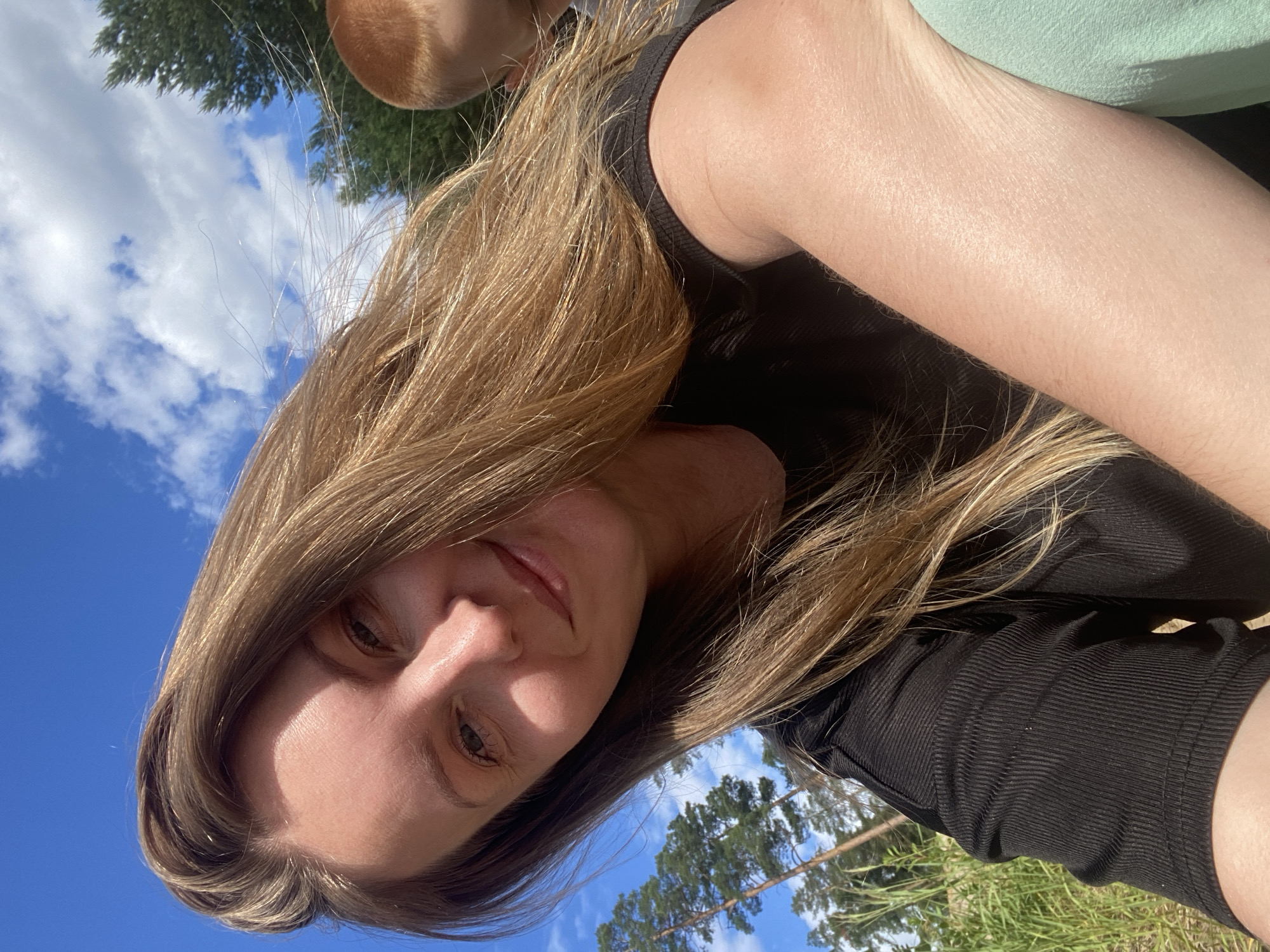 A young woman with long, blonde hair is taking a selfie outdoors on a sunny day. She is wearing a black t-shirt and light green pants. Her right arm is visible, resting in front of her. In the background, there are trees and a blue sky with white clouds. There is also a glimpse of another person, a child with red hair, to the right of the woman. The environment appears to be a natural, possibly wooded area, which could be within or near the city of Novyi, Russia.