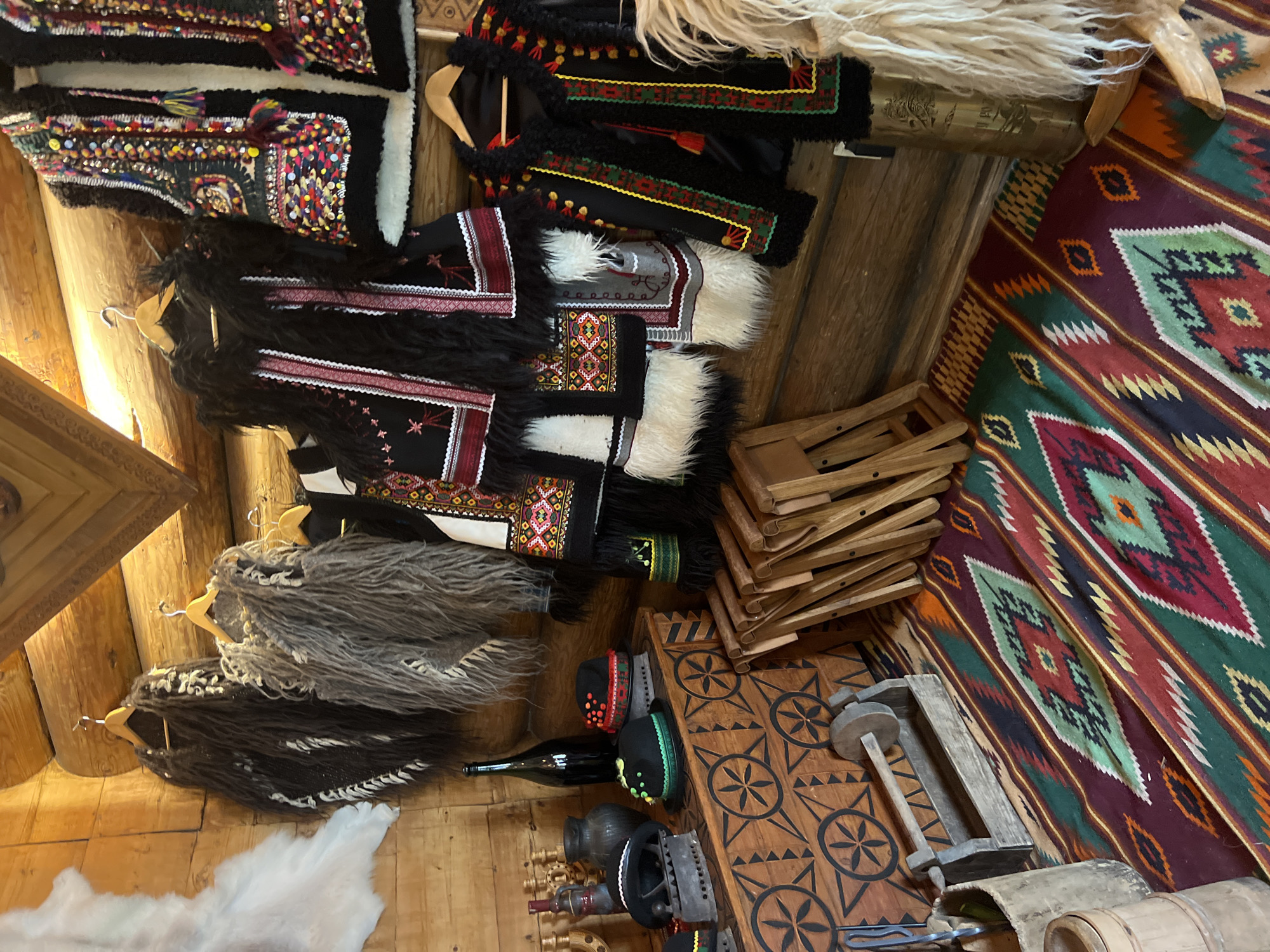 The image depicts a display of traditional Hutsul clothing and artifacts within a wooden interior, likely in Yaremche, Ukraine. On the wooden walls hang several vests and fur garments, characterized by intricate embroidery, beadwork, and fur trim. In the foreground, a wooden chest with carved circular motifs is visible, adorned with several hats and a vintage iron. To the right, a collection of wooden chairs are stacked, and a richly patterned rug with geometric designs covers the floor. The overall impression is one of a cultural exhibition or a shop showcasing traditional Hutsul craftsmanship.