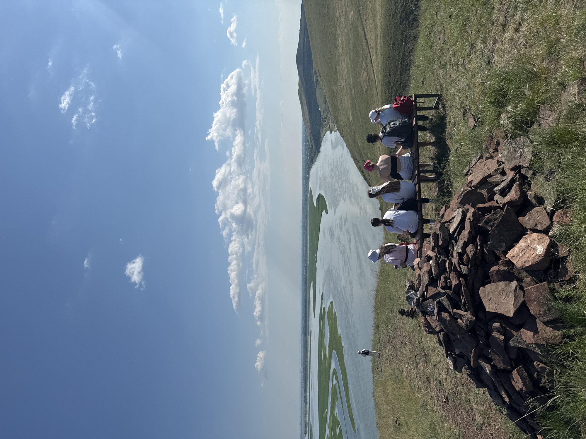 A group of six individuals, five seated on a bench and one standing in the distance, are positioned on a grassy hillside overlooking a wide river. The seated individuals are facing away from the camera, their backs to the viewer. They appear to be young women, some wearing hats and backpacks. In the foreground, a large pile of reddish-brown rocks is stacked on the grass. The river, with a meandering channel and lush green banks, dominates the mid-ground. The background shows rolling, grassy hills and a hazy sky with scattered clouds. The scene suggests a recreational outing or hike in a natural setting. The geographical context indicates this view is from the vicinity of Ust'-Abakan, Russia, likely overlooking the Abakan River or a similar waterway in the region.