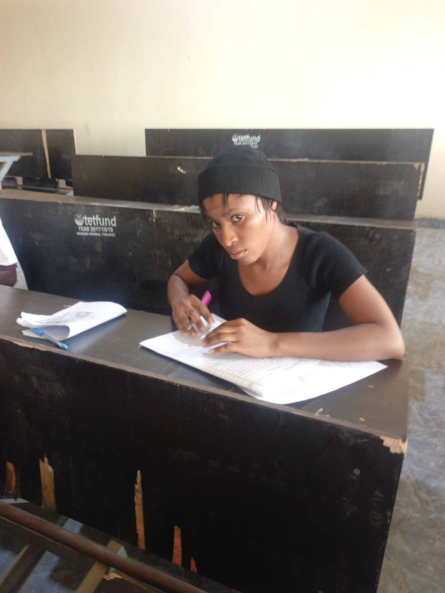 A young woman is seated at a desk in what appears to be a classroom or lecture hall. She is wearing a black beanie and a black short-sleeved top. The woman is looking towards the camera. She is writing on a piece of paper with a pink pen. Additional papers are on the desk to her left, alongside a blue pen.
The desk is part of a row of fixed seating, and the word "tetfund" is printed on the back of the seating. The location may be the city of Akwanga, Nigeria. The desks appear worn. The floor is plain and unmarked.
