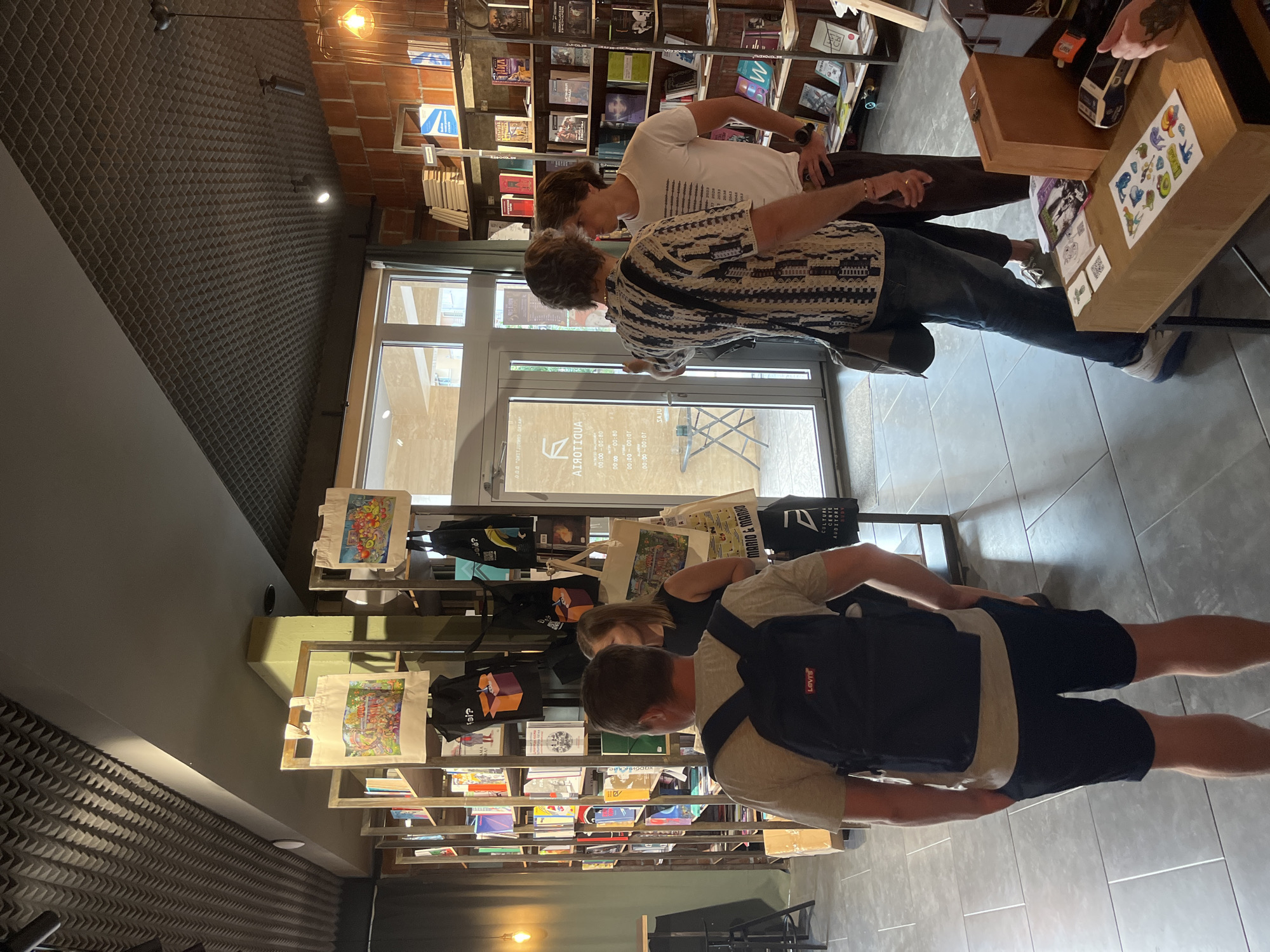 The image depicts the interior of a bookstore or cultural center. Several people are visible. In the foreground, a man wearing a grey t-shirt, dark shorts, and a black backpack is standing with his back to the camera, looking towards a bookshelf. To his right, a woman in a black dress is talking to another person. Further into the scene, two men are interacting, with one wearing a patterned shirt and jeans, and the other in a white t-shirt and dark pants. The environment includes shelves stocked with books, some tote bags hanging on display, and a wooden counter with various items on it, including stickers and a small wooden box. The lighting suggests an indoor setting, possibly with natural light coming from a large window or glass door visible in the background. The walls are partly exposed brick, and the ceiling has a textured, sound-dampening material.