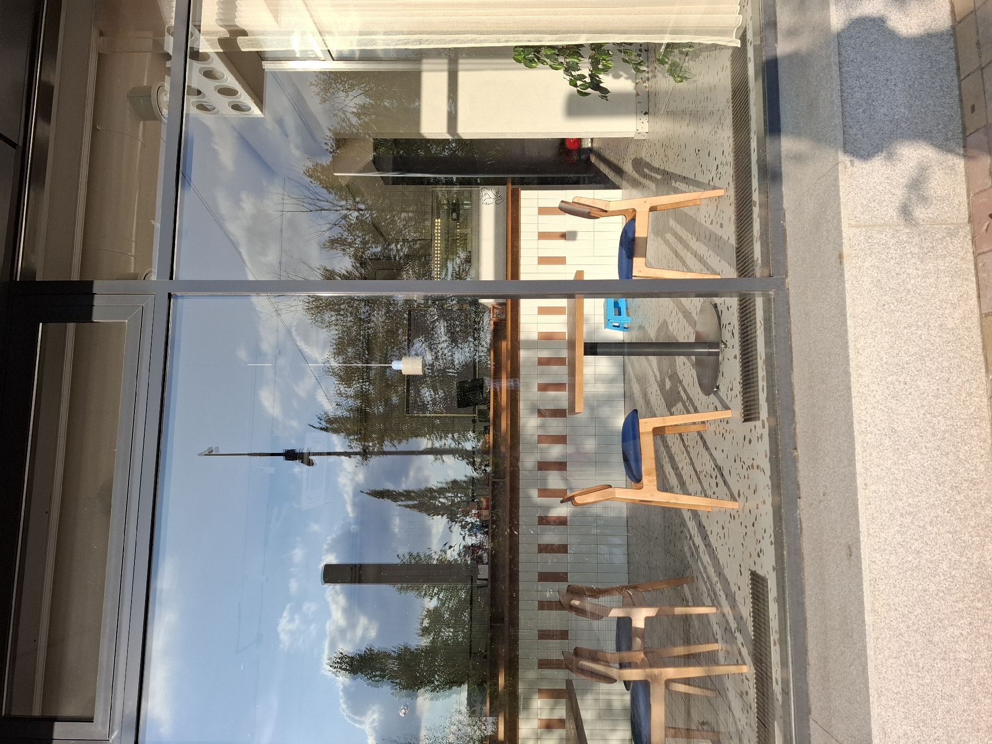 The image shows the exterior of a modern cafe or restaurant in Lisakovsk, Kazakhstan, as seen through a large glass window. The reflection in the glass dominates the scene, showing a bright blue sky with scattered clouds and the silhouettes of trees and industrial structures, possibly a tall chimney or tower. Inside the cafe, there are several wooden chairs with blue cushions arranged around tables. A counter area with tiled walls is visible in the background, suggesting a food or drink service area. A white curtain hangs to the right, and a potted plant is placed near the window. The floor inside appears to be tiled with a speckled pattern. A shadow of a person is cast on the pavement outside the window in the foreground. The lighting suggests it is late afternoon.