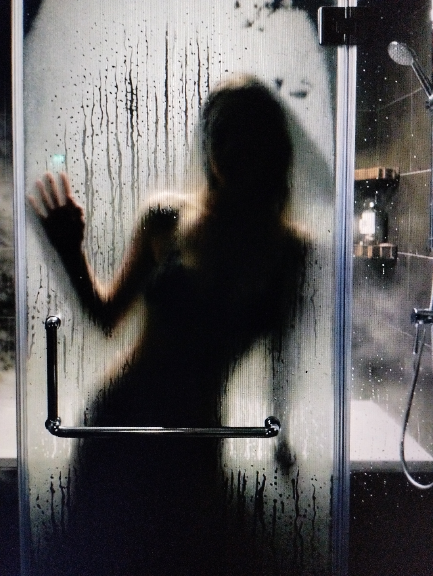 A silhouetted figure is obscured behind a steamy shower door in a bathroom. The glass is covered in condensation, with water droplets and streaks running down its surface. The figure appears to be leaning against the door, with one hand pressed against the glass. Their form is indistinct, appearing as a dark shadow. Visible elements of the bathroom include a silver showerhead and a portion of a bathtub. The lighting is dim, contributing to the shadowy and obscured nature of the scene.