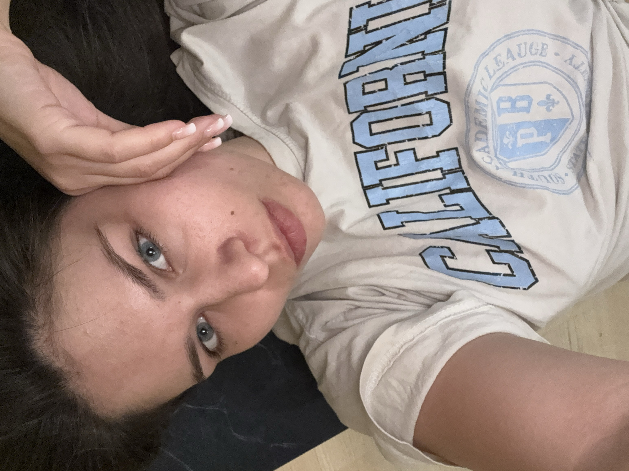 This is a close-up, high-angle selfie of a young woman lying down, with her head tilted to the side. She has dark hair and striking blue eyes. Her right hand is raised, resting gently on her forehead and cheek, with manicured fingernails visible. She is wearing a light-colored t-shirt with light blue text and a crest on it. The text on the t-shirt reads "CALIFORNIA" in large, blocky letters, and the crest contains the words "ACADEMIC LEAUGE" and what appears to be an insignia with letters. The lighting suggests it is likely indoors, possibly in a bedroom or living area, with soft ambient light. The overall mood is relaxed and introspective.
