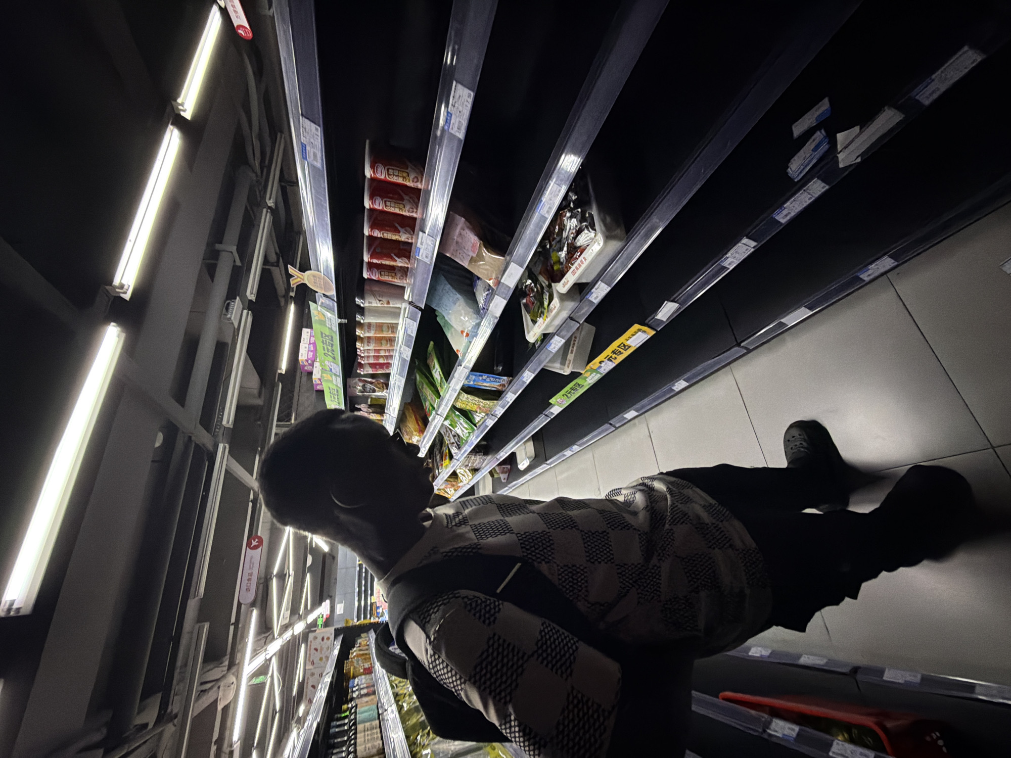 In a dimly lit supermarket aisle in Jiangjin, China, a young Black man with dark skin, wearing a patterned shirt and dark pants, stands with his back to the camera. He is looking down at a shelf filled with various packaged goods. The shelves are dark, and some are sparsely populated. To the man's left, there are more shelves stocked with bottled beverages and other items. The ceiling is equipped with fluorescent lighting, some of which are illuminated. The floor is tiled in a light gray color. The overall atmosphere suggests a quiet shopping experience.