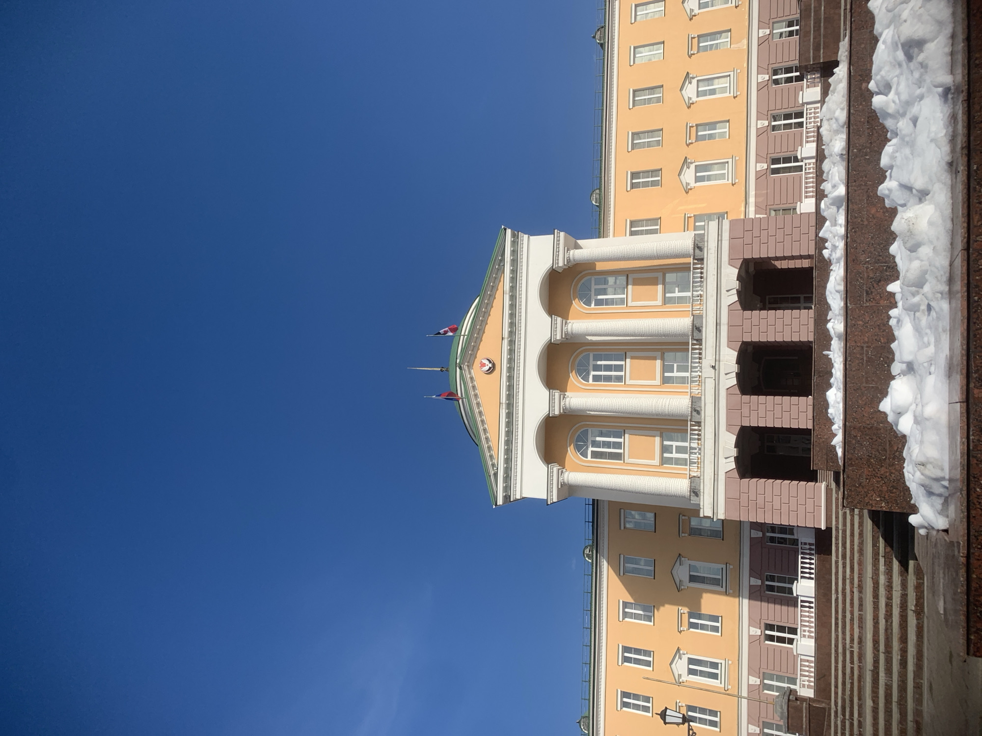 The image depicts the House of the Government of the Udmurt Republic located in Izhevsk, Russia. The multi-story building features a pale yellow facade with white window frames and trim, under a clear, bright blue sky. Its central section is characterized by a prominent portico with four large, fluted white columns supporting a white pediment. Above the pediment, a green domed roof is visible, topped by two flagpoles. The flag of the Russian Federation (white, blue, red horizontal stripes) and the flag of the Udmurt Republic (black, white, red vertical stripes) are flown from these poles.
Below the main portico, the ground floor features a reddish-brown stone base with arched entranceways. Numerous windows, predominantly rectangular with white sashes, are distributed across the facade, while the central upper story includes arched windows. White balustrades are present on the central balcony and along lower sections of the building. In the foreground, a series of broad, reddish-brown granite steps leads up towards the building. Accumulations of dirty white snow are visible on these steps and on granite ledges, suggesting a winter or early spring setting. Strong sunlight illuminates the scene, casting clear shadows on the building and ground elements. No individuals or discernible actions are present in the frame.