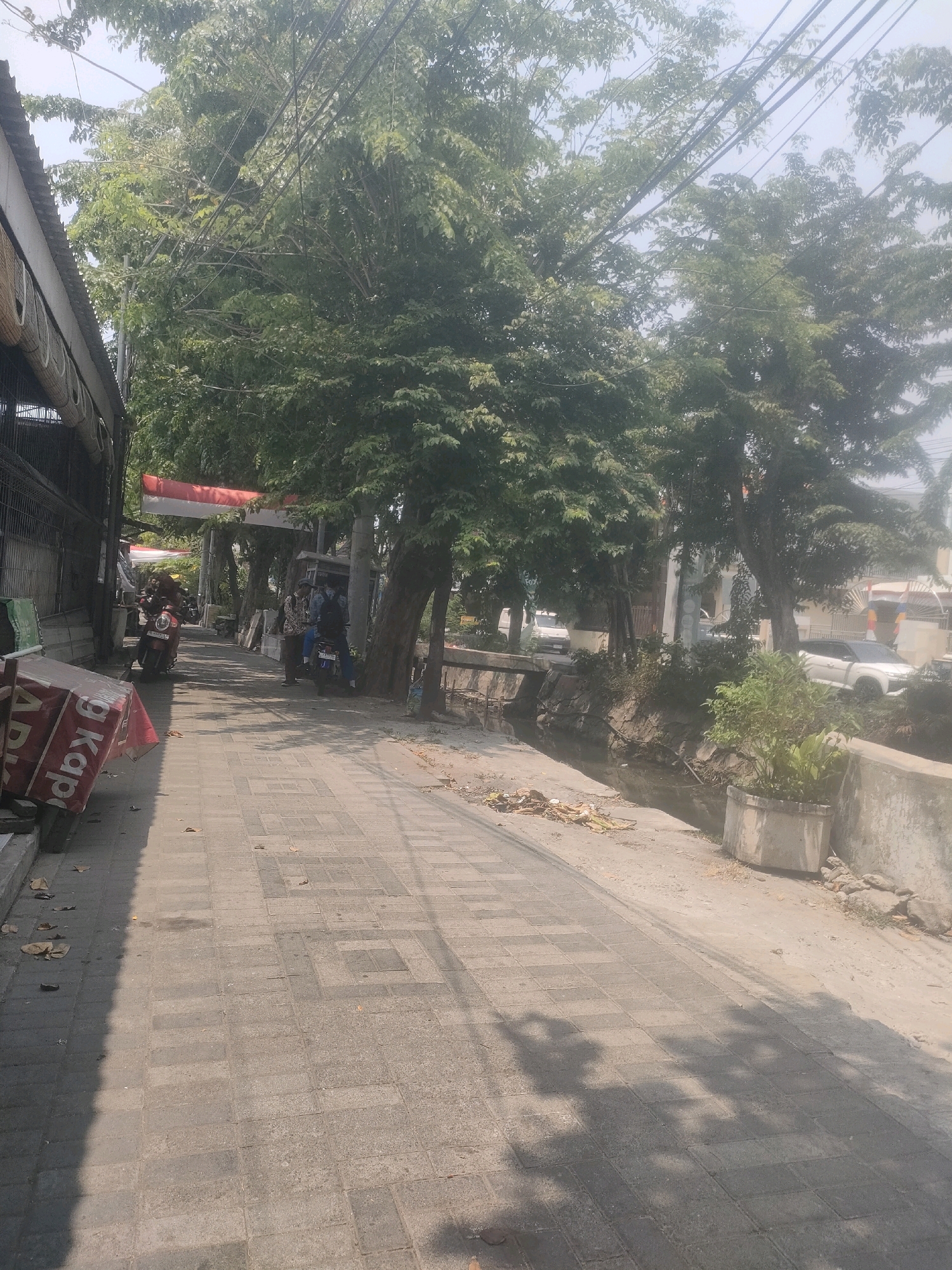 This image depicts a street scene in Surabaya, Indonesia, on a bright, sunny day. A paved sidewalk, laid with square gray tiles, occupies the foreground and mid-ground, running alongside a narrow, shallow canal. The canal is lined with rough stone on one side and a concrete barrier on the other, with some vegetation growing within and around it. A large planter with green plants sits on the concrete barrier.
Along the left side of the sidewalk, there is a building with a metal grate and a sloped roof. In front of this building, a red and white sign, partially obscured by a tarp, leans against a low wall. Further down the sidewalk, two motorcycles are parked, one red and one blue. On the blue motorcycle, two young men are seated, facing away from the camera.
Behind the sidewalk and canal, a row of mature trees with dense green foliage provides shade. An Indonesian flag, red and white, is suspended between poles above the street. In the background, a white car is visible near a building with a white facade. The overall impression is one of an active, everyday street in a tropical urban environment.