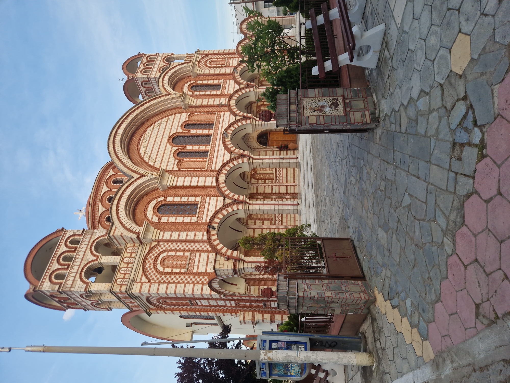 The image captures a street scene in Ασπροβάλτα, Greece, focusing on the Church of Saint George. The church is a multi-storied building with a mix of red brick and white stone, constructed with distinct arched windows and entrances. A bell tower stands to the left side of the church, topped with a cross. The sidewalk in front of the church is paved with a variety of colored stones. A metal fence, featuring a cross emblem, encloses a small garden area with plants. A park bench stands alongside the fence. The sky is clear and blue. In the lower foreground is a utility pole, and a damaged public telephone booth. There are no people visible in the frame.