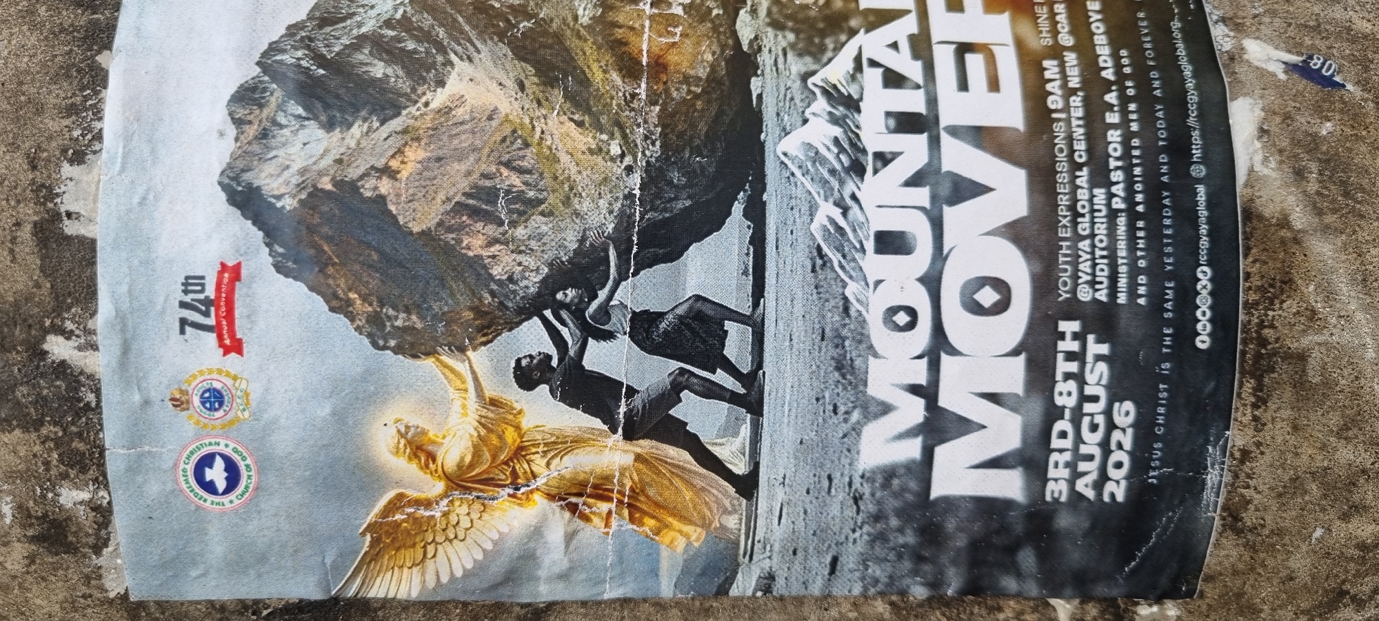 A poster is displayed on a rough, textured concrete surface. The poster itself appears aged and somewhat damaged with visible rips and creases. The imagery on the poster depicts a golden-winged angel with outstretched arms supporting a large boulder. Beside the angel, a man and a woman are also pushing against the boulder, appearing to exert significant effort. The background features a mountainous landscape. At the top of the poster, two circular logos are visible, one with a dove symbol and the text "THE REDEEMED CHRISTIAN CHURCH OF GOD" and another circular logo with a crown and stars. The text "74th Annual Convention" is also present at the top. Below the main illustration, large text reads "MOUNTAIN MOVER." Further down, details such as "3RD-8TH AUGUST 2026," "YOUTH EXPRESSIONS | 9AM," "@YAYA GLOBAL CENTER, NEW AUDITORIUM," and "MINISTERING: PASTOR E.A. ADEBOYE AND OTHER ANOINTED MEN OF GOD" are printed. A Bible verse, "JESUS CHRIST IS THE SAME YESTERDAY AND TODAY AND FOREVER," is also visible. Social media handles and a website address, "rccgyaglobal" and "https://rccgyaglobal.org," are at the bottom of the poster. The poster is likely a promotional material for an event at the Redeemed Christian Church of God. The presence of the "YAYA GLOBAL CENTER" suggests a location within the Redeemed Christian Church of God's network, and while specific city location is not explicitly stated on the poster, the Redeemed Christian Church of God has a significant presence in Ikeja, Nigeria, where its headquarters and international convention center are located.