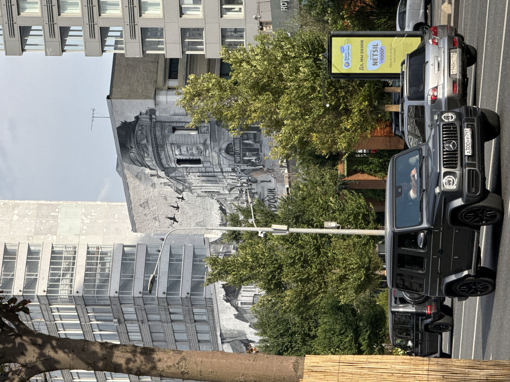 This is a street-level view of a mural depicting a historical urban scene on the side of a building. In the foreground, a dark grey Mercedes-Benz G-Class is driving on a paved road, its license plate reading "A300YP 797". Behind it, another similar black Mercedes-Benz G-Class is also in motion. Further back, a silver Mitsubishi Outlander is visible with the license plate "T140KC 199". Other vehicles and trees line the street, and a bright yellow billboard with Russian text is present on the right side of the image. The mural itself is rendered in grayscale and features architectural elements, figures, and what appears to be text in a historical font. Tall modern buildings and trees form the backdrop of the scene. The mural is located on a building at the intersection of Tverskaya Street and Sadovaya-Triumfalnaya Street in Moscow, Russia.