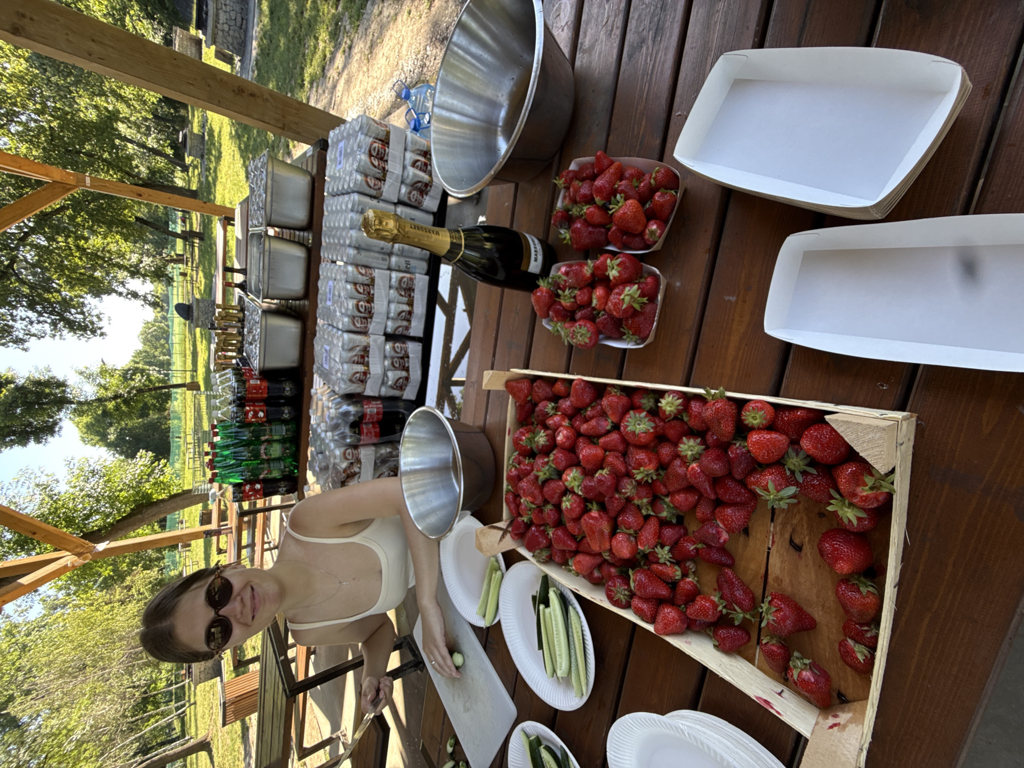 A woman wearing sunglasses and a white top is seen cutting cucumbers on a white cutting board at a wooden table in an outdoor setting. The table is laden with a variety of items: a large wooden box filled with strawberries, two smaller containers also filled with strawberries, a bottle of champagne, several stacks of canned beverages, numerous plastic bottles of soda, a large metal bowl, stacks of white paper plates, and two rectangular white trays. The scene appears to be set up for an outdoor event or gathering. In the background, trees and open grassy areas suggest a park or recreational area in the Cukarica Urban Municipality, Serbia. The wooden table is part of a wooden structure, possibly a picnic shelter.