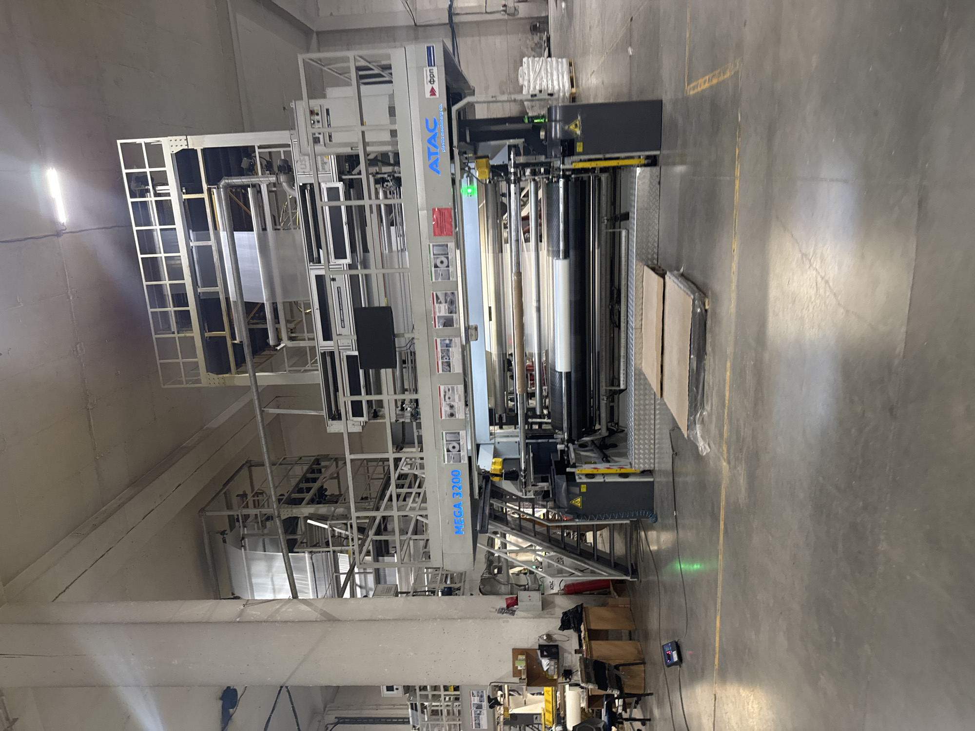 The image shows a large industrial machine, an ATAC MEGA 3200, installed in a factory or warehouse setting. The machine is complex, featuring multiple levels and components including rollers and platforms. The location is implied to be a factory or warehouse due to the presence of a large, unfinished concrete floor, structural support beams, and exposed utilities in the ceiling. It's also implied the location is in Perm, Russia, due to the "APAC" logo on the machine. There are no people visible in the image. The environment is brightly lit with a mix of natural and artificial light. Several industrial components such as boxes, cables, and metal plates are scattered on the floor.
