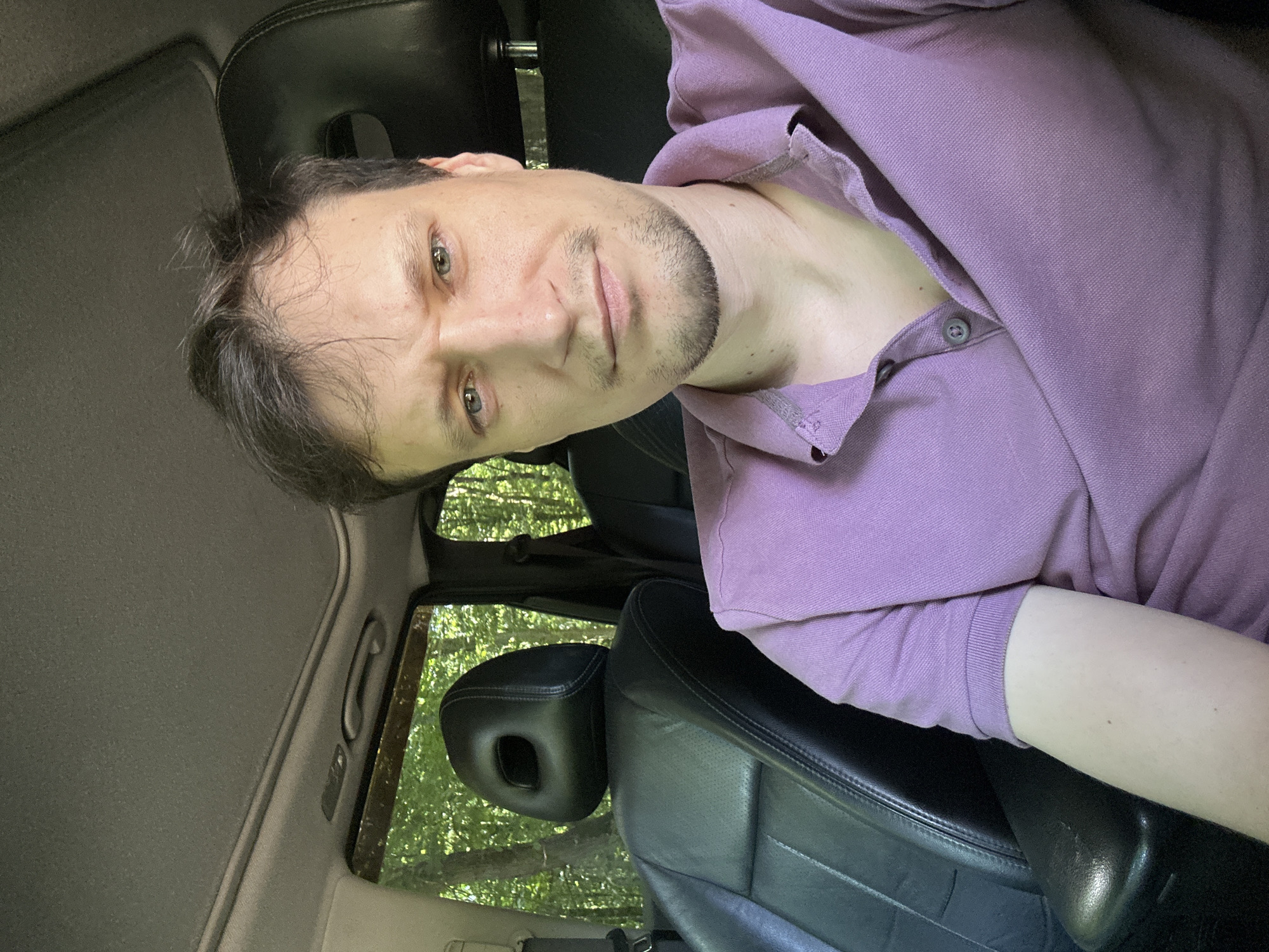 A man with light skin, short brown hair, and light blue eyes is seated in the driver's seat of a car. He is wearing a purple polo shirt. The interior of the car appears to be black leather. Outside the car, trees and greenery are visible through the windows, suggesting an outdoor, possibly wooded, location. The car has a sunroof, which is open. The image is a selfie, taken from a slightly low angle. The man has a slight stubble on his chin and a faint goatee. The man's left shoulder and arm are visible in the foreground, resting on the center console. The overall lighting suggests daytime.