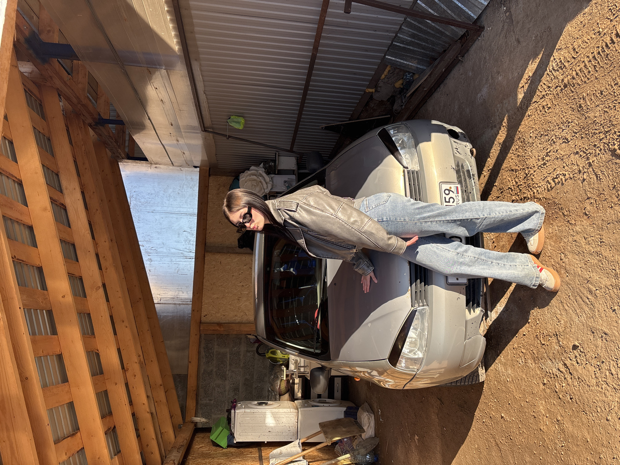 The image depicts a young woman posing on the hood of a light-colored car within an open-sided garage or carport, likely in Фоки, Russia. The setting features a rustic, makeshift structure with exposed wooden beams, corrugated metal roofing, and a dirt ground.
The woman is lying face down, stretched out on the car's hood, with her arms extended and hands resting flat. She is wearing a grey or brown leather jacket, light blue jeans, and dark sunglasses. Her dark hair flows around her head.
The car is a silver or beige crossover-style vehicle, possibly a Nissan, with some visible wear on its front bumper. A Russian license plate is prominently displayed on the front, reading "159 RUS".
The surrounding area includes various stored items, such as two old washing machines, shovels, plastic bottles, and a green bucket, suggesting a utility or storage space. One side of the structure has a wooden lattice wall, while the other side features concrete blocks and wooden paneling.
Bright sunlight illuminates the scene, casting strong shadows on the ground and indicating a clear, sunny day, likely morning or late afternoon given the shadow angles. The overall atmosphere is casual and relaxed, capturing a posed moment.