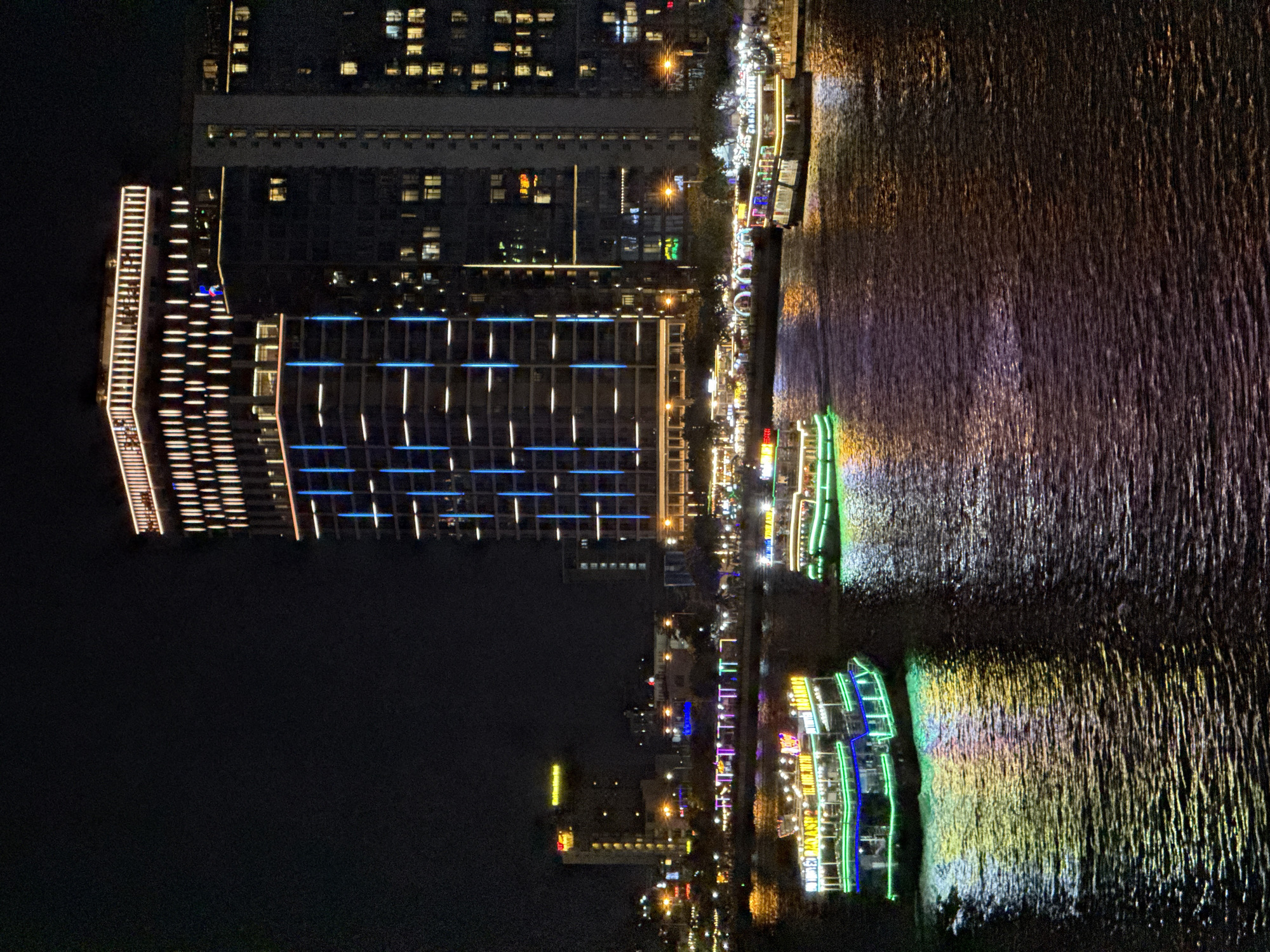 This is a nighttime, eye-level, medium shot photograph taken from a slight elevation, capturing a scenic view of the Han River in Da Nang, Vietnam. The river dominates the foreground, its dark waters reflecting the vibrant lights of the city. Two illuminated tour boats are navigating the river. The boat on the left is larger and more prominent, adorned with bright green, blue, and yellow lights, casting colorful reflections on the water. The boat on the right is smaller and further away, also lit with white and yellow lights, its reflection visible on the rippling surface.
In the background, the city skyline is illuminated by a multitude of lights. Dominating the right side of the frame is a tall, modern skyscraper with a distinctive architectural design, featuring vertical lines of blue and white lights and a top section outlined in warm yellow light. To the left of this skyscraper, other buildings are visible, some with illuminated windows and rooftop lights. The riverbank is lined with a series of lights, suggesting a promenade or entertainment area. The overall scene conveys a lively and bustling atmosphere of a vibrant city at night.