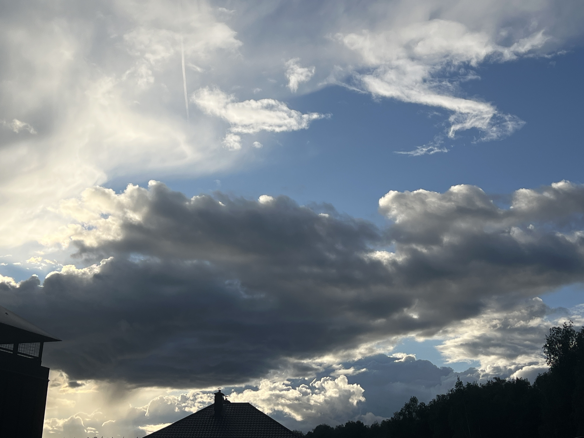 The image depicts a cloudy sky above a residential area in the city of Unknown, Unknown. The sky is a mix of bright blue and various shades of white and dark gray clouds. There is a thin contrail from an aircraft visible in the upper left part of the image. The lower portion of the image features the silhouettes of buildings, a house roof with a chimney, and trees against the bright sky. There are no people visible in the image.