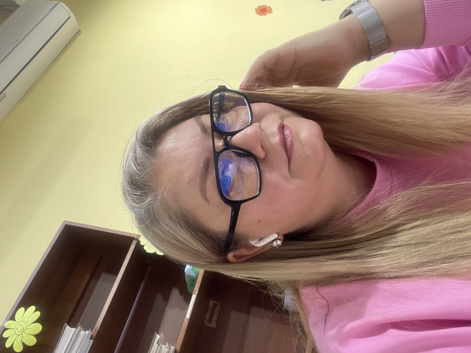 A selfie taken indoors shows a woman with blonde hair and wearing glasses, a pink sweatshirt, and a silver watch. She is touching her hair with her hand. There is a shelf with files in the background, and an air conditioner is visible in the upper left corner. A yellow wall is also visible. There is no information that confirms the location of the picture is in the city of Izhevsk, Russia.