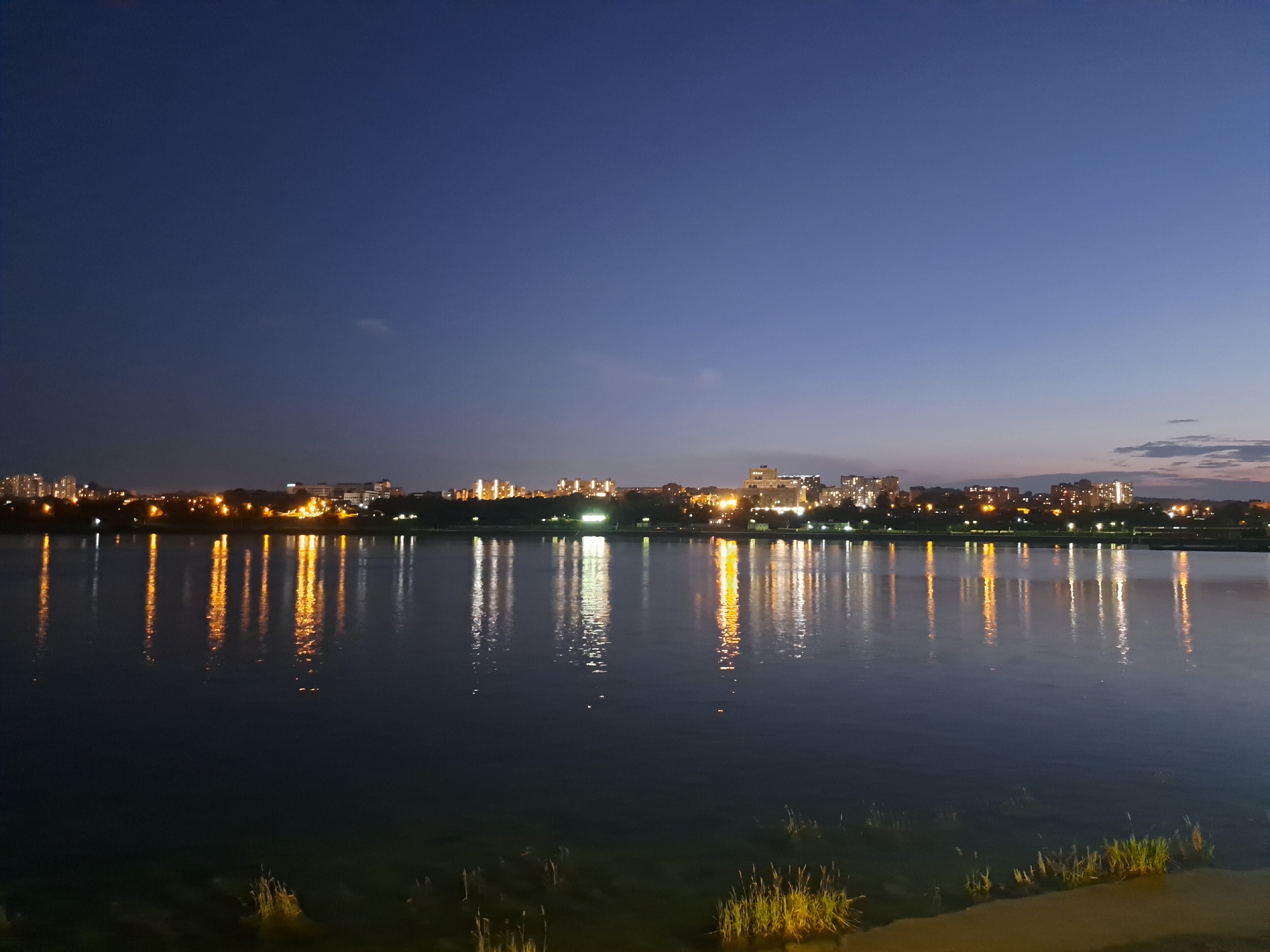 A twilight view of a city skyline across a body of water, likely a river or lake, is captured in this image. The sky transitions from a deep blue at the top to a lighter hue near the horizon. A faint, wispy cloud is visible in the upper portion of the sky. The city, on the opposite bank, is illuminated by numerous lights, indicating it is evening or nighttime. Buildings of various heights are discernible, with their lights reflecting in the water. The reflections create elongated streaks of yellow and white light on the dark surface of the water. In the foreground, the water is calm, with some reeds or grass growing at the very edge, illuminated by the ambient light. The scene suggests a peaceful urban waterfront setting.