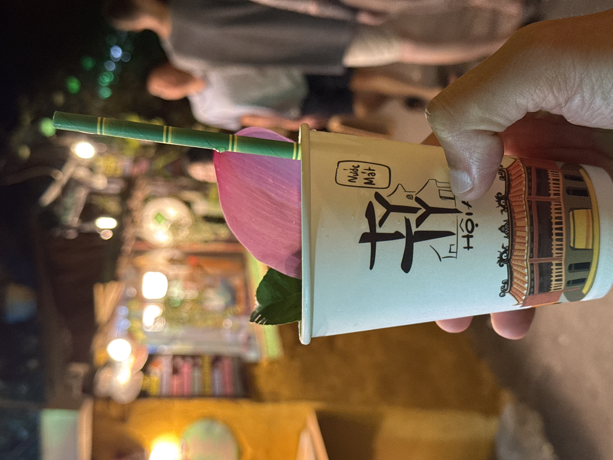 A right hand holds a light-colored paper cup, prominently displaying black stylized Vietnamese text including "Nước Mát" within a rectangular border and an illustration of a traditional Vietnamese building with an ornate tiled roof. Inside the cup, a single pink lotus petal, a green leaf, and a green bamboo-patterned paper straw with yellow stripes are visible; a small water droplet clings to the straw near the rim. The scene is illuminated by numerous out-of-focus artificial light sources, creating a bokeh effect against a dark background, indicating nighttime. In the blurred mid-ground to the right, at least two indistinct human figures are discernible, suggesting a populated public area such as a night market or street in Hội An Ward, Vietnam.