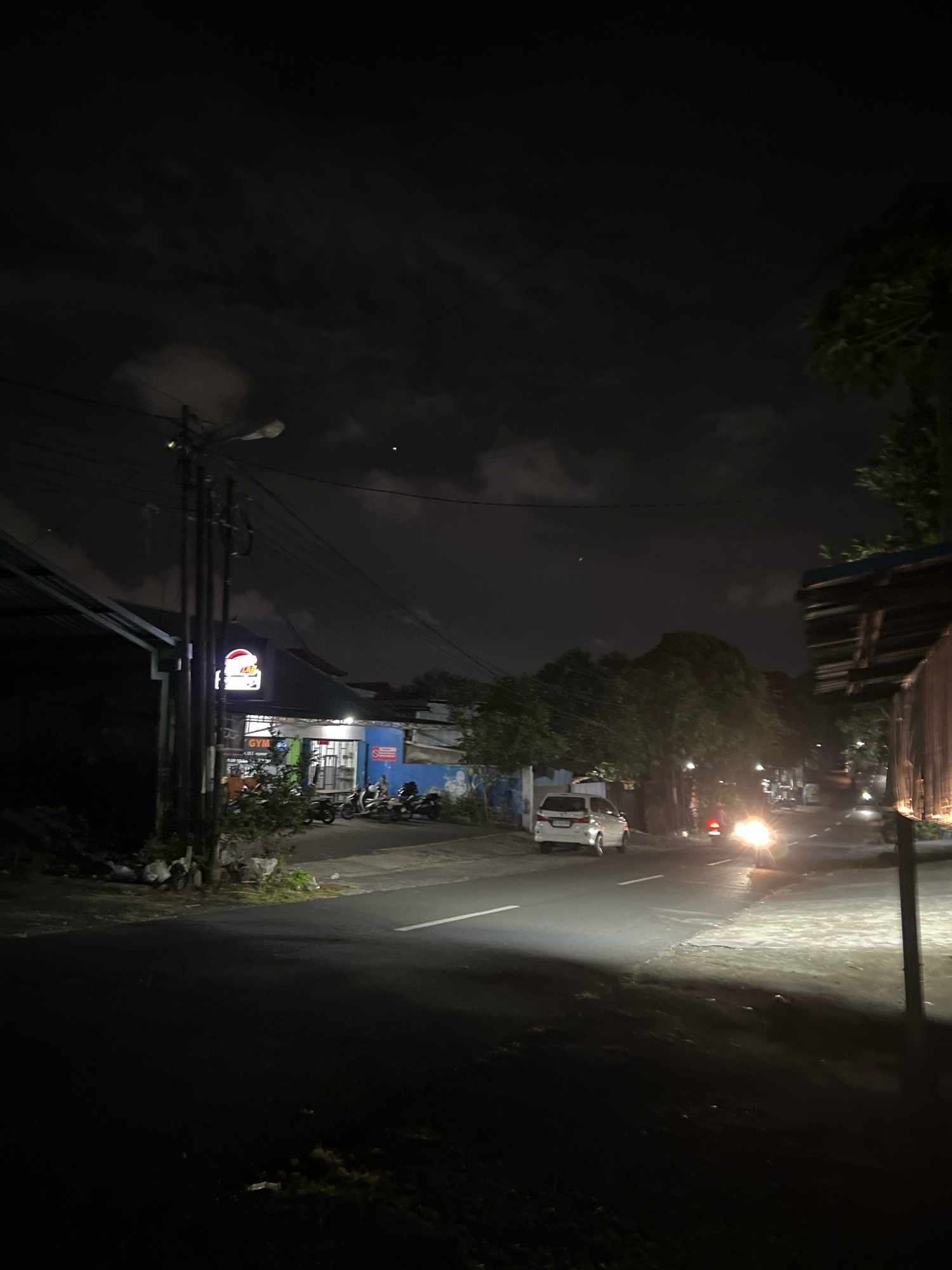 The image depicts a street scene at night in Denpasar, Indonesia. A white van is driving on the road, with its headlights illuminating the scene. There are several motorcycles parked along the side of the road, and a few people are visible near a building with a sign that says "GYM". The street is paved, and there are trees and buildings lining the road. The sky is dark with some clouds.