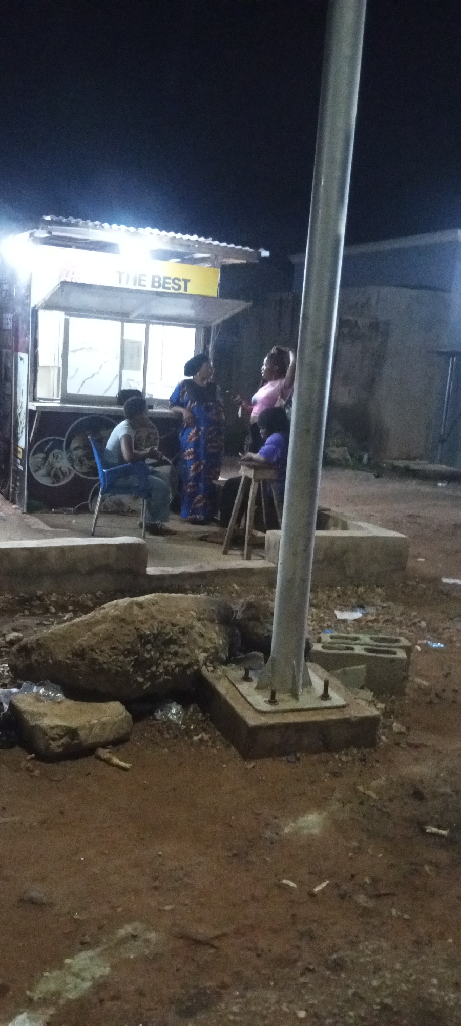 The image depicts an outdoor scene at night in Adankolo, Nigeria, near a food stall. A silver pole is prominently positioned in the foreground, anchored to a concrete base with bolts. To the left, a food stall with a corrugated metal roof is illuminated by interior lights. A yellow sign above the stall reads "THE BEST".
Four individuals are visible around the stall. One person, seated on a blue chair, is wearing a light-colored top and dark pants. Another person, in a blue and patterned dress, stands to the right of the stall. Two other individuals are engaged in conversation near the stall; one wears a pink top and the other a purple top. A wooden stool is visible near the stall, and a large rock lies on the ground in the foreground, along with scattered debris. The ground appears to be dirt and gravel. The overall environment suggests a commercial area at night.