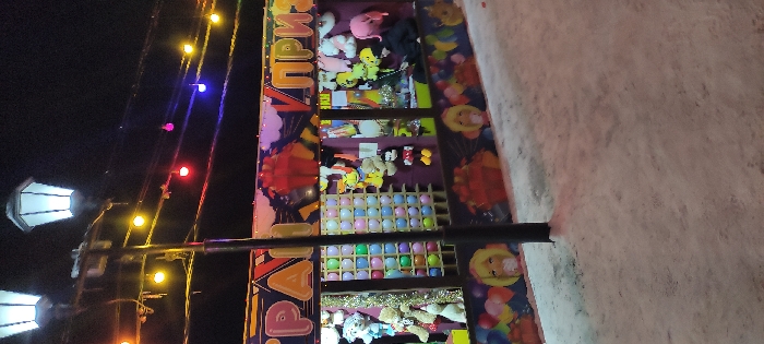 An outdoor carnival game is viewed in the evening. It features rows of prizes including stuffed animals and toys. Below, there are sections of balloons in various colors arranged in a grid. The game structure is decorated with colorful designs. A lamp post with three lights stands nearby. String lights are also visible above, adding to the festive atmosphere. The ground appears to be covered in snow.