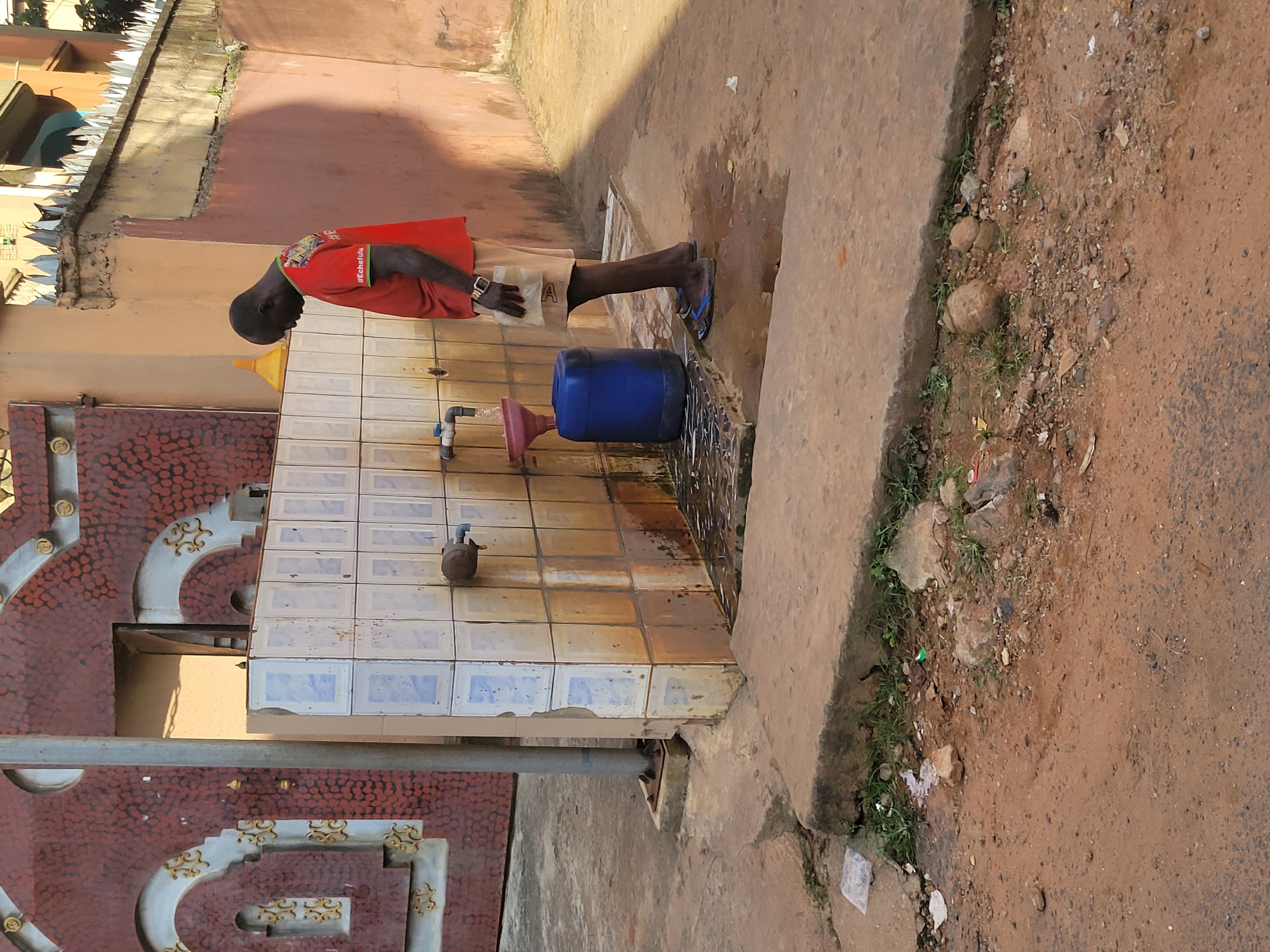 In Onitsha, Nigeria, a man wearing a red t-shirt with the hashtag #Echefula printed on it and light-colored shorts stands at an outdoor public faucet. He is holding a small white object, possibly a bar of soap. The faucet is attached to a tiled wall, which shows signs of wear and staining. Below the faucet, a pink funnel directs water into a large blue plastic container. The ground around the faucet is wet and appears to be a mix of dirt and concrete, with some debris scattered about, including plastic bags and rocks. In the background, a large decorative red gate with ornate gold detailing is visible, along with a section of a building. The scene suggests a practical, everyday activity taking place in a somewhat weathered urban environment.