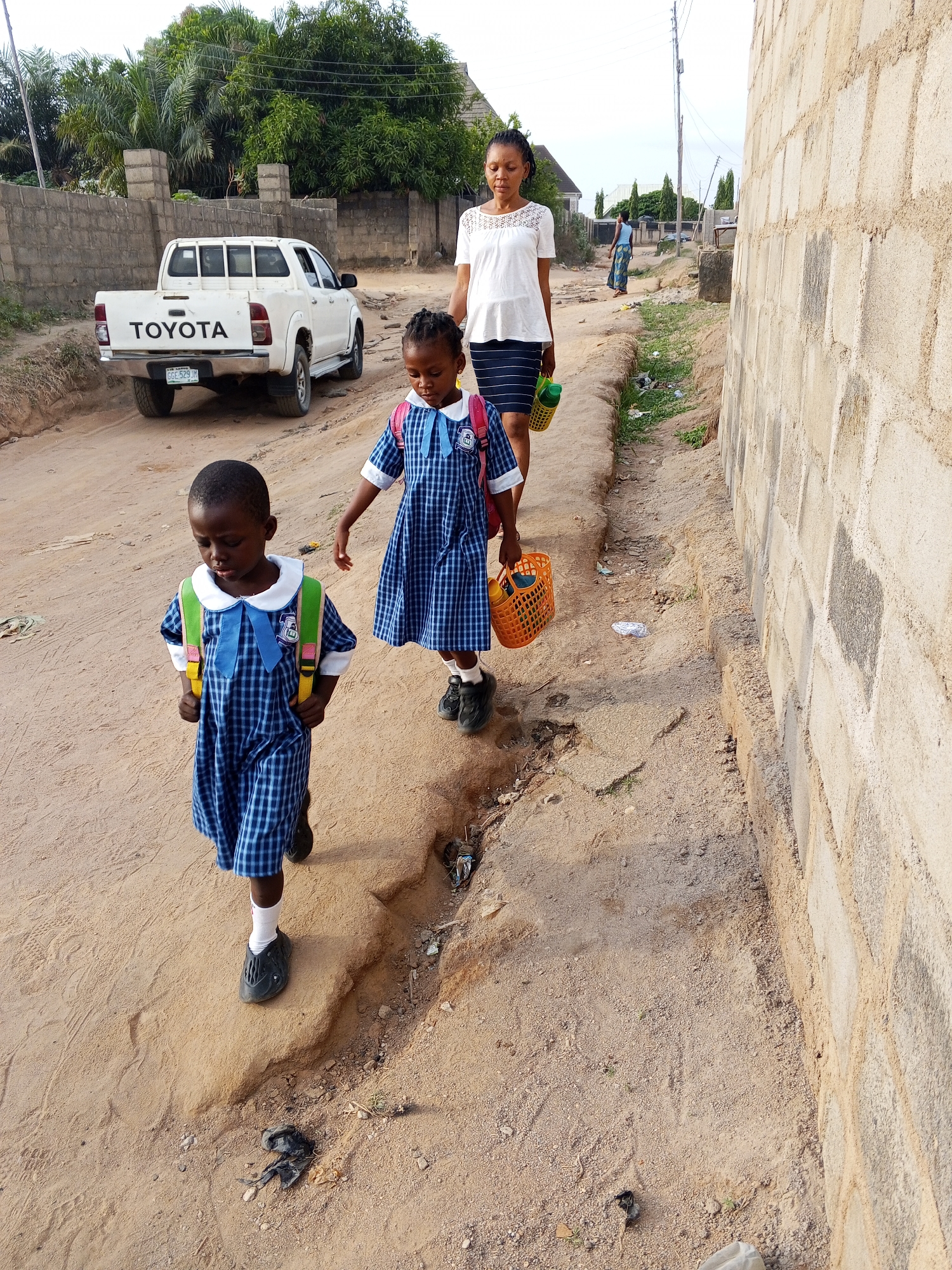 In Adonkolo, Nigeria, a woman and two young children, all Black, walk along a dirt road beside a concrete block wall. The children wear matching blue plaid school uniforms with backpacks and carry lunch baskets. The woman wears a white blouse and striped skirt. A white Toyota pickup truck is parked on the left side of the road. The road is uneven and littered with debris. In the background, other buildings and vegetation are visible. A utility pole is visible in the distance.