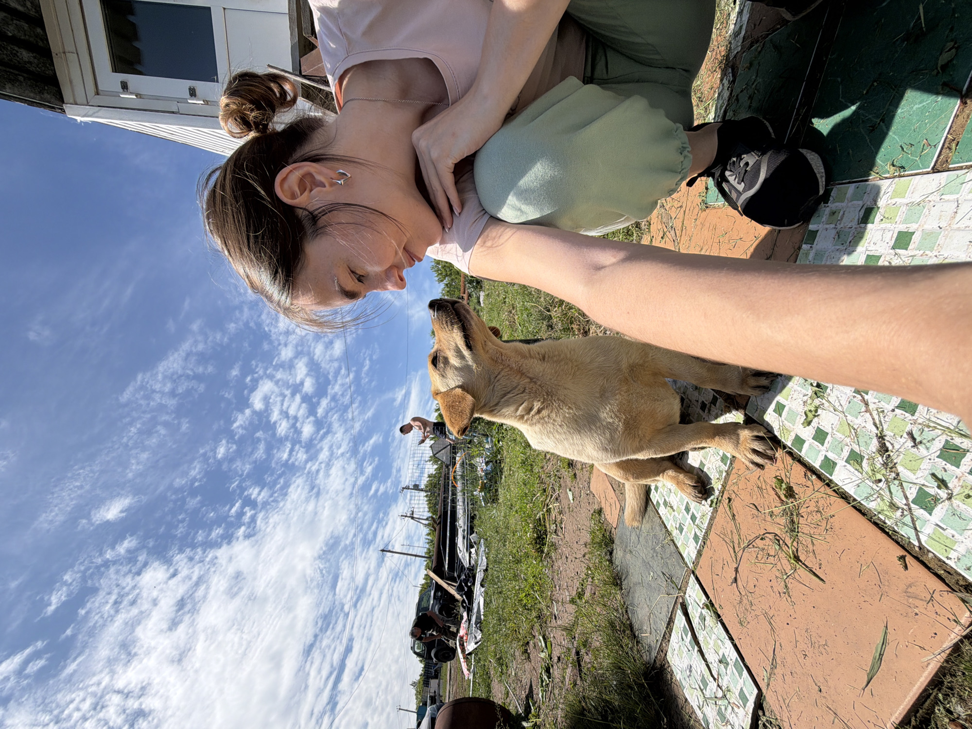 This image, captured in Sarapul, Russia, depicts an outdoor scene under a bright blue sky with scattered cirrocumulus clouds. In the foreground, a young adult female with brown hair styled in a messy bun, wearing a light pink t-shirt, light green sweatpants, and black sneakers, crouches on a tiled surface. Her face shows a gentle expression as she looks towards a light brown, short-haired dog. The dog sits attentively on the tiles, looking up at the woman with its ears perked, establishing a direct interaction. The woman's right forearm is extended towards the camera. The ground consists of dirt patches, sparse green grass, and patterned square tiles in shades of green, brown, and white, with scattered organic debris. In the mid-ground, a dark-colored SUV or jeep is visible near piles of what appear to be disassembled structures, metal frames, and tarps. Two adult males are present in the background; one is shirtless and standing while looking at a mobile phone, and the other is bending near the vehicle. A white, single-story building with a dark roof is partially visible in the far right background, with utility poles and wires against the sky. The scene is illuminated by strong natural sunlight, casting distinct shadows.