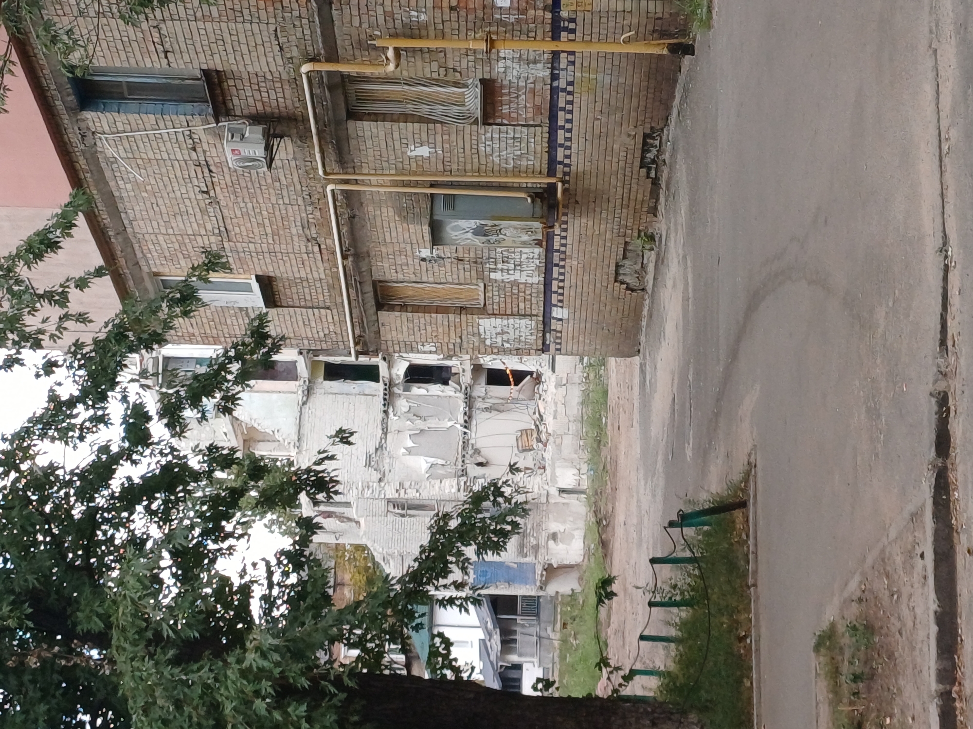 The image shows a street scene in Kyiv, Ukraine, with a focus on two adjacent buildings. The building on the right is made of brick, with visible yellow pipes running along its facade. One window on this building has metal bars. An air conditioning unit is mounted on the brick wall to the left of a window. The building on the left appears to be under demolition, with exposed interiors and debris visible. The street in the foreground is paved, with a curb and some grass and trees to the left. The overall impression is one of urban decay and reconstruction.