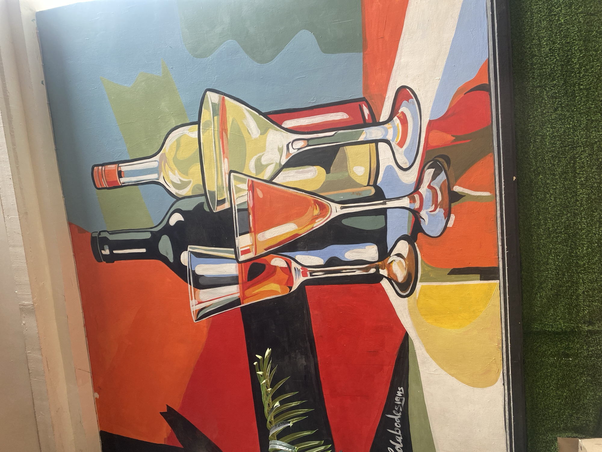This is a photograph of a mural painted on a wall. The mural depicts a still life of two bottles and four glasses filled with various colored liquids. The style of the mural is abstract and cubist, with bold geometric shapes and colors. The bottles are dark green and pale yellow. The glasses are clear and filled with amber and yellow liquids, and some appear to have ice or garnishes.
The background of the mural is a mosaic of colorful geometric shapes in shades of red, orange, blue, green, and yellow. There is a black and red striped pattern behind the bottles and glasses. To the left of the bottles and glasses, there is a green leafy sprig. In the bottom left corner of the mural, there is a signature that reads "Colabodesigns 1995".
The mural is framed by a dark border and appears to be located indoors, possibly in a restaurant or bar. Below the mural is a textured surface that resembles artificial turf. The lighting suggests the image was taken during the day, with some sunlight possibly coming from an unseen window. The location is specified as Howayi, Nigeria, but this information is not visually verifiable from the photograph itself.
